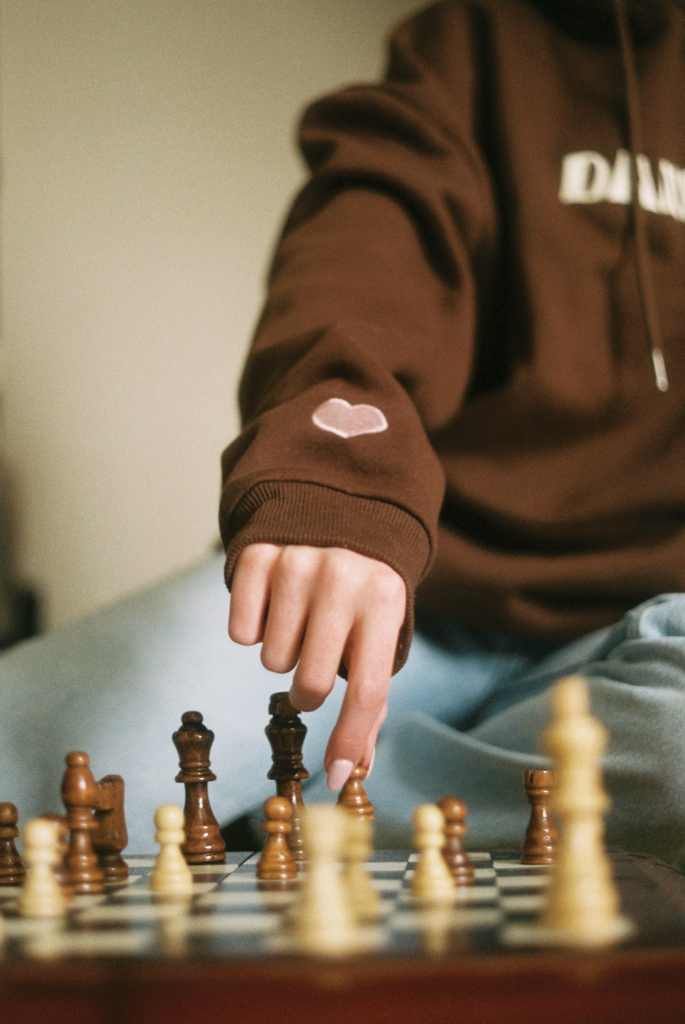Woman plays chess in brown hoodie with small pink heart on sleeve and DANDY text on chest.