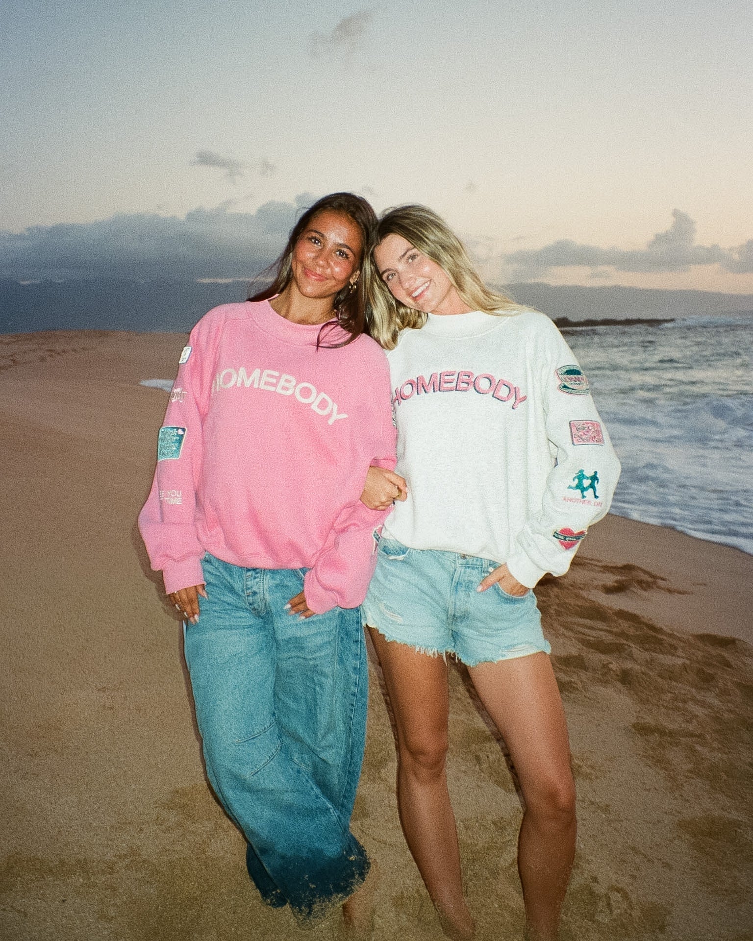 Two people wearing 'Homebody' sweatshirts on a beach with ocean and sky in the background.
