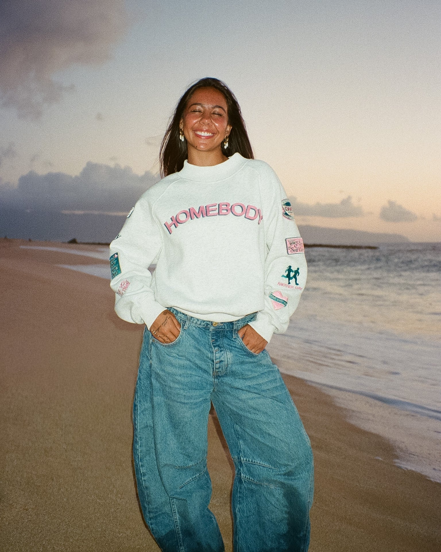 Person wearing a 'Homebody' sweatshirt on a beach at sunset