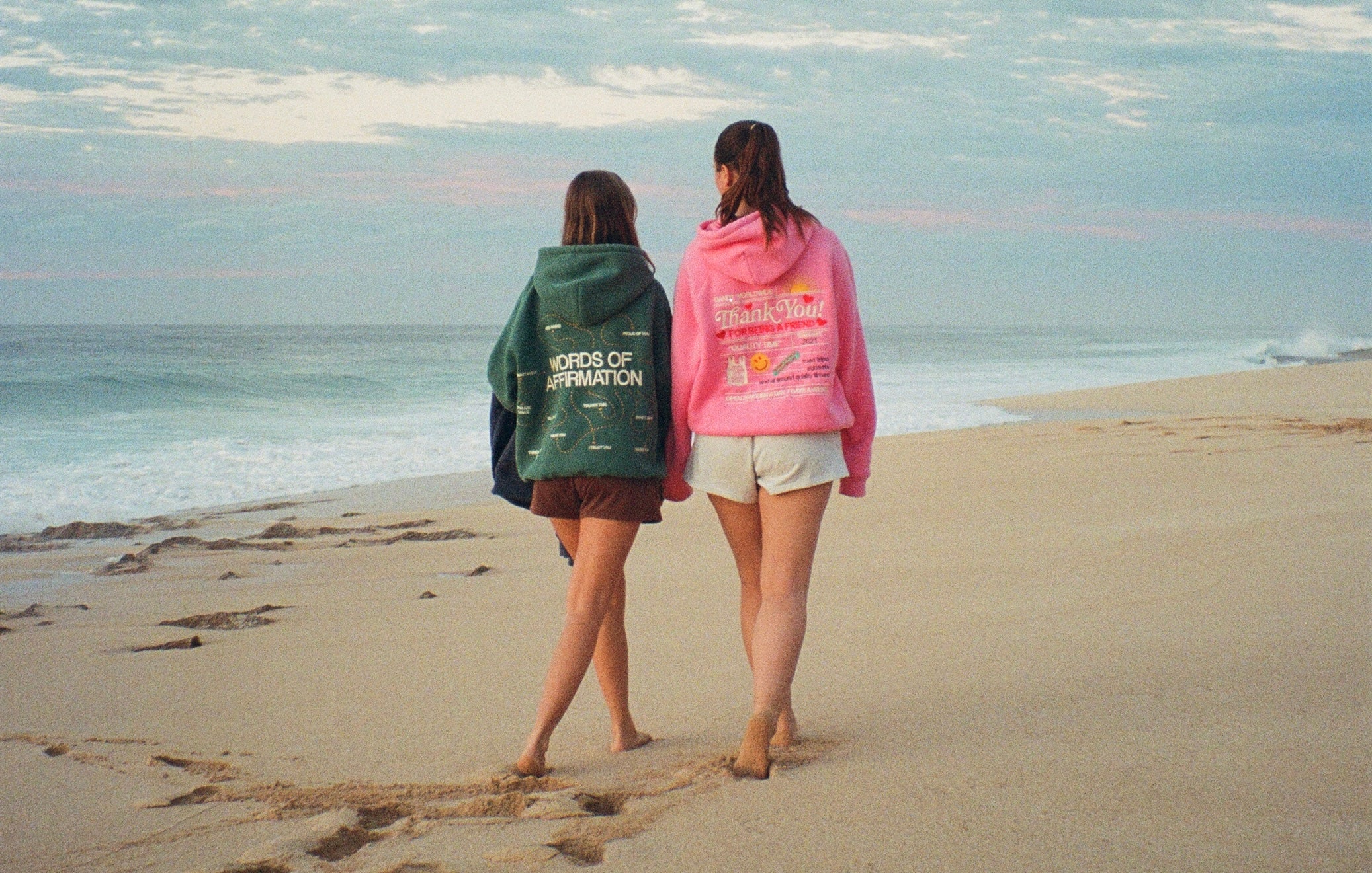 Two women walk barefoot along the beach wearing Dandy hoodies—
one green, one pink—leaving footprints in the sand