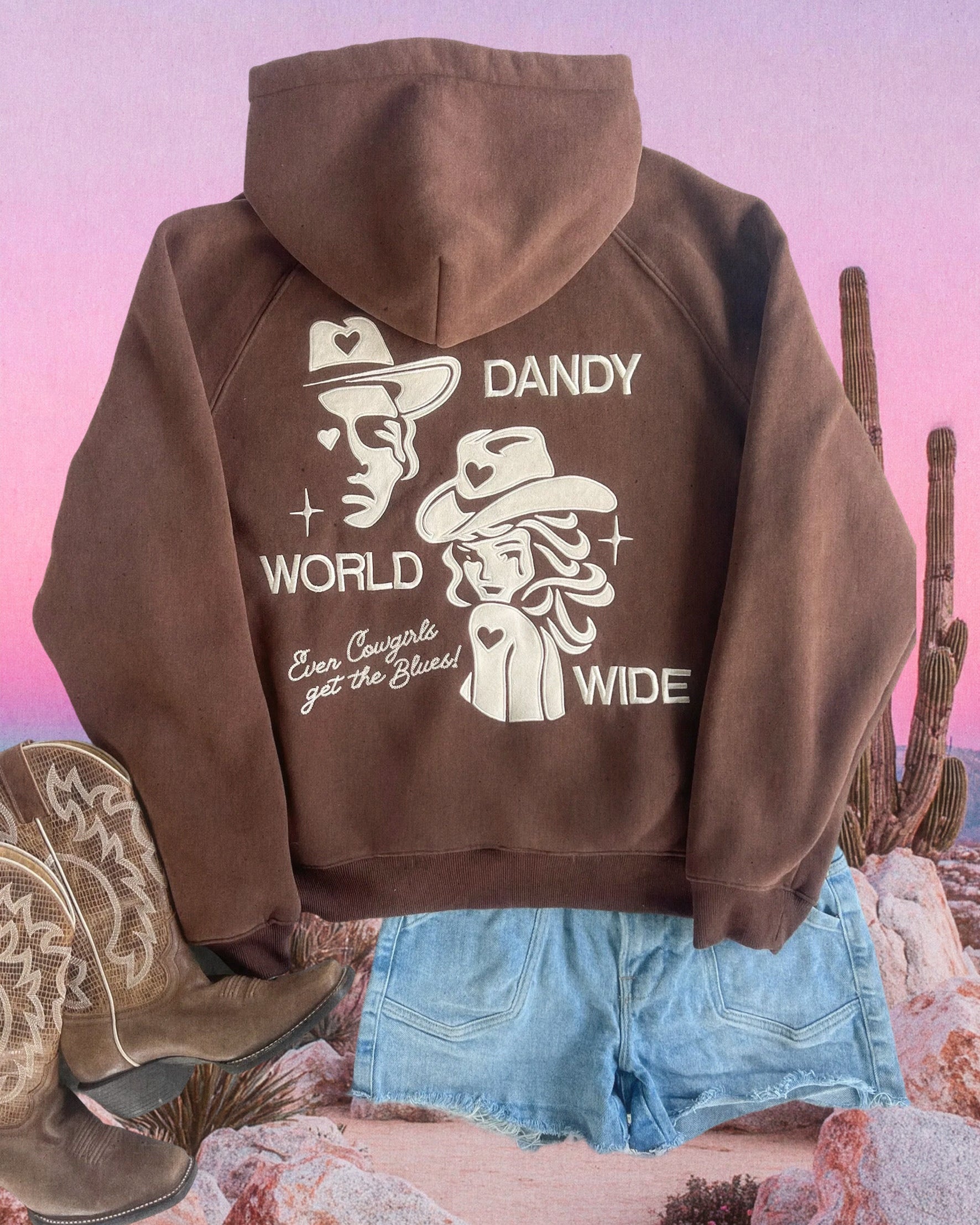 Brown hoodie with graphic design on the back, worn with denim shorts and cowboy boots, against a desert landscape.