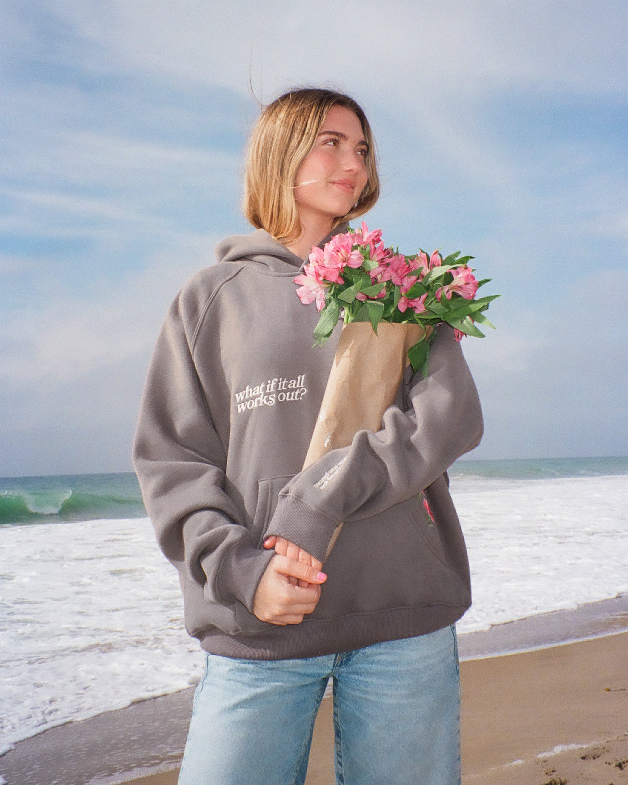 Person wearing a hoodie with text on a beach holding flowers