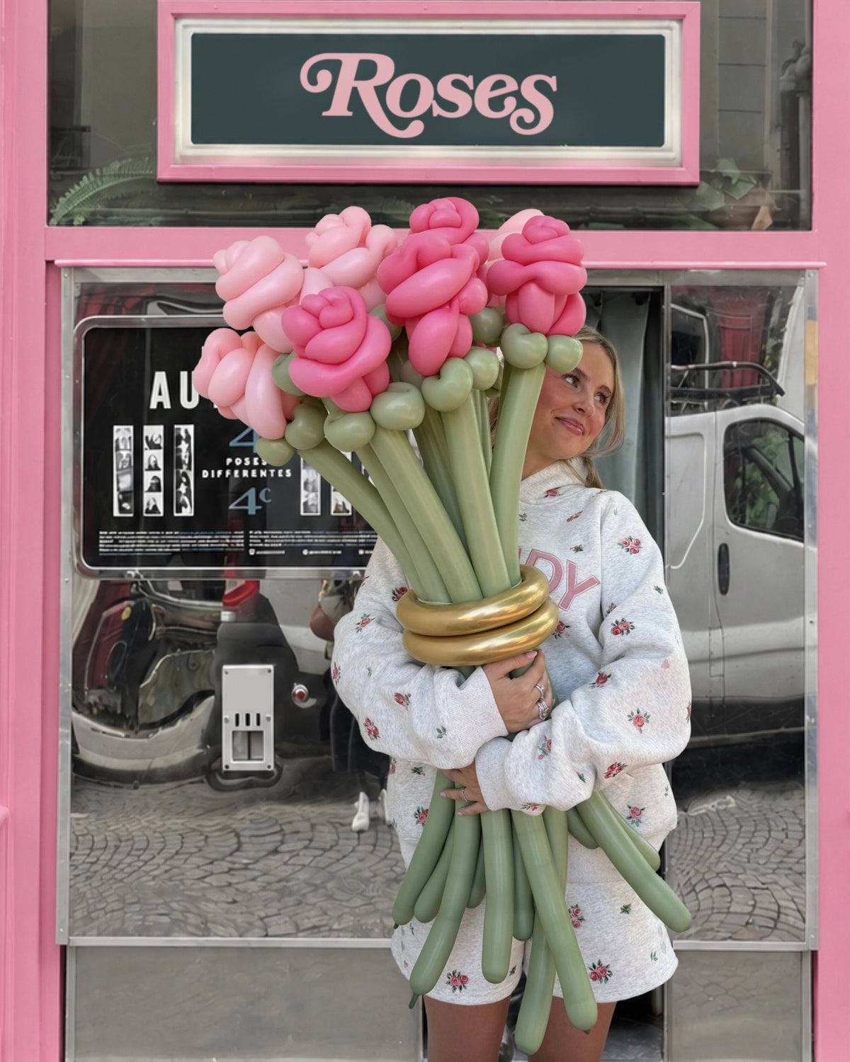 A woman stands in front of a pink storefront window holding a large bouquet made of pink and light green balloon roses with a metallic gold band around the stems. The shop sign above reads 'Roses'.