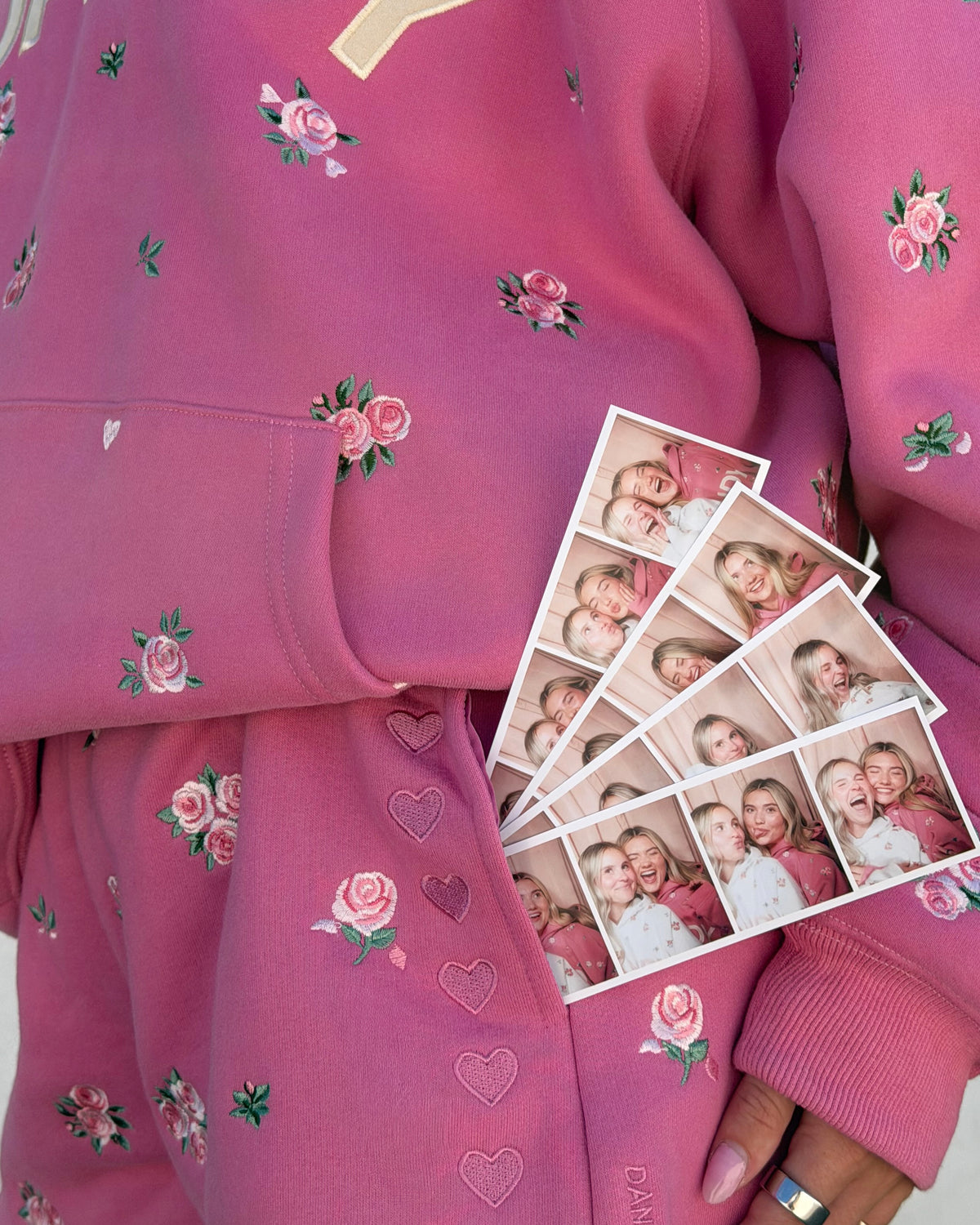 A close-up of a person wearing a pink sweatshirt and matching sweatpants with embroidered pink roses, green leaves, and heart motifs. The person is holding a fan of five printed photo strips showing a smiling couple.