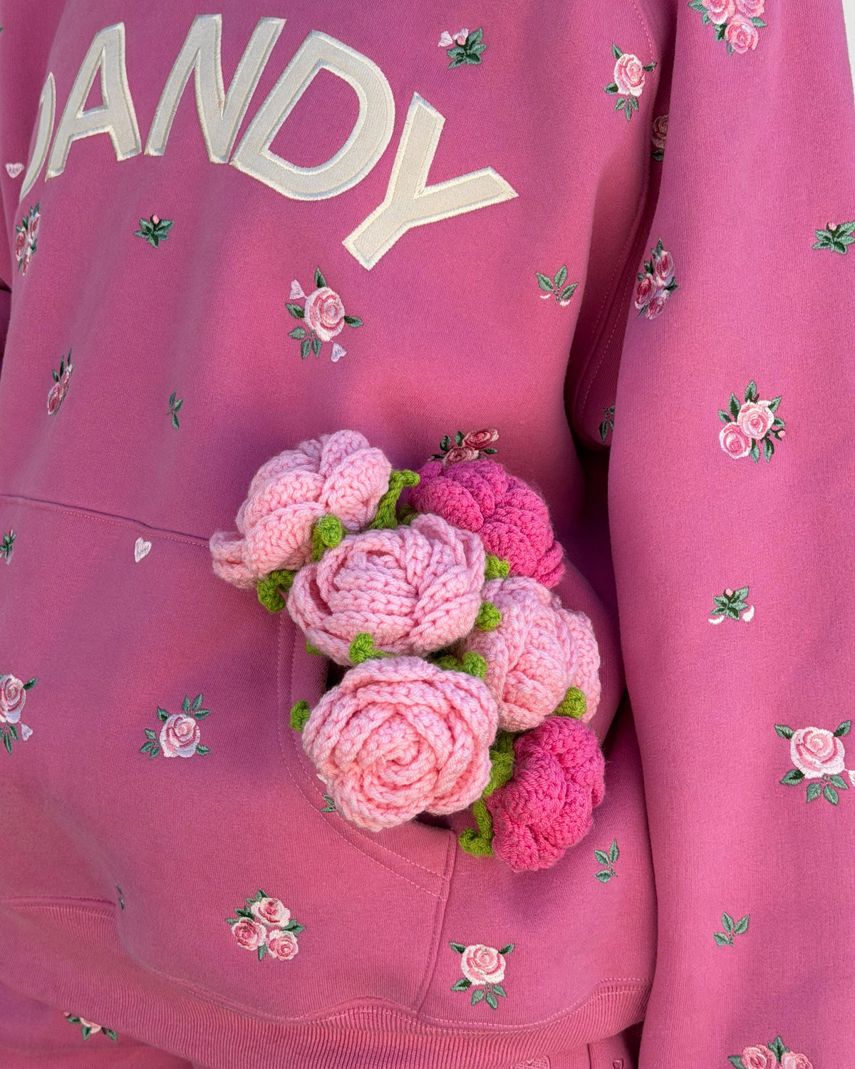 A close-up of a pink hoodie featuring embroidered pink roses, a kangaroo pocket, and the partial text 'NANDY' on the chest. A small pink and green crocheted rose bouquet is tucked into the pocket.