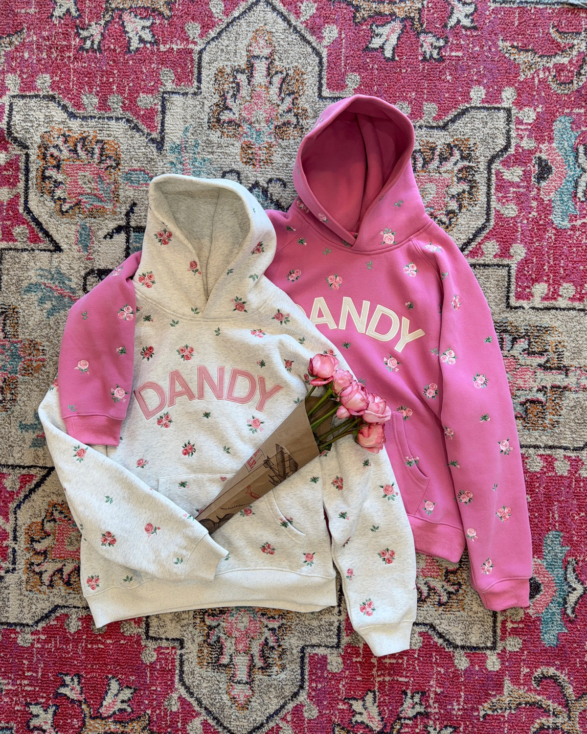 Two floral hoodies laid flat on a patterned rug: one light cream and one bright pink, both with small pink flowers and the word 'DANDY' across the chest. A bouquet of pink flowers rests over the hoodies.