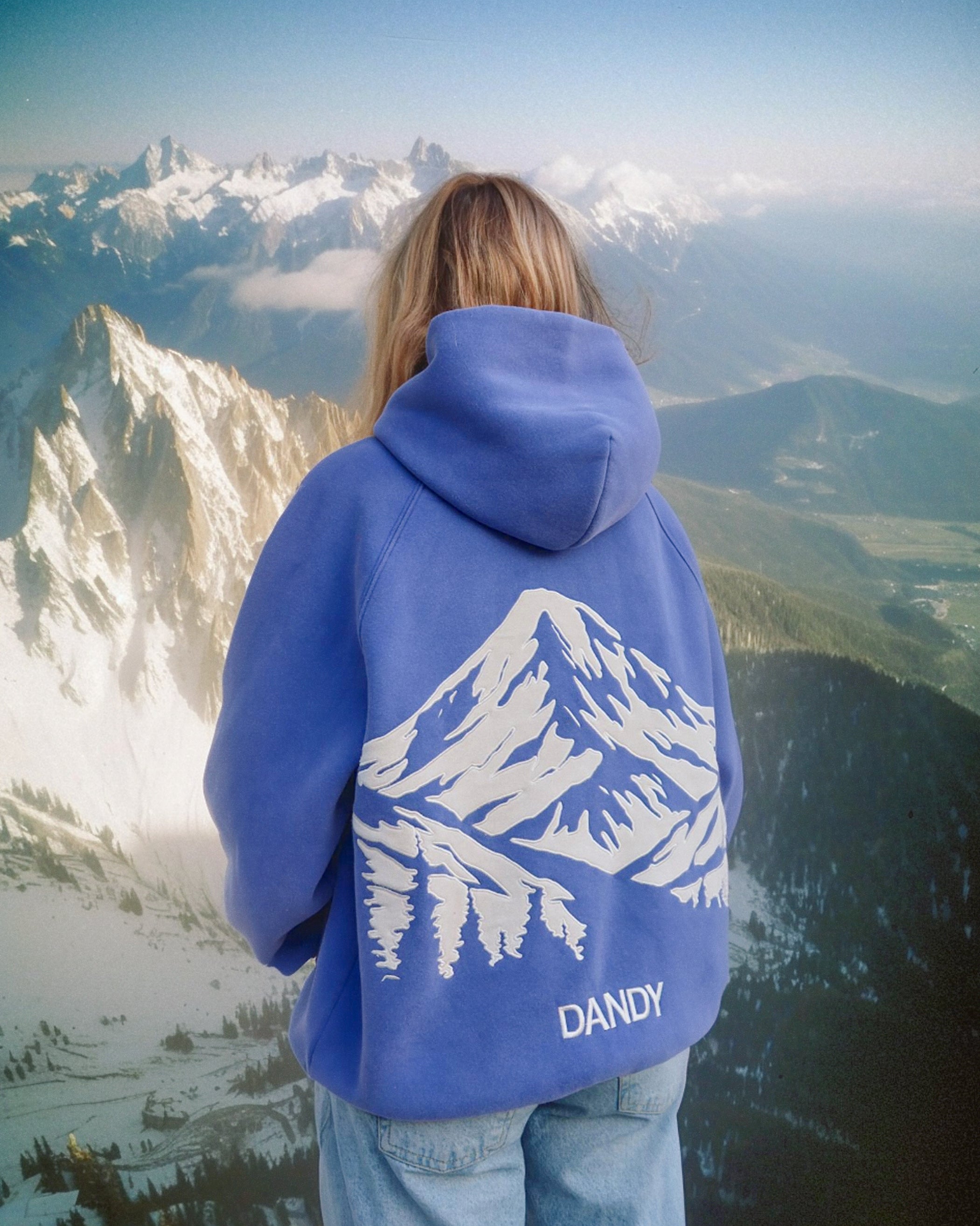 Person wearing a blue hoodie with mountain design and 'DANDY' text, standing on a mountain peak.