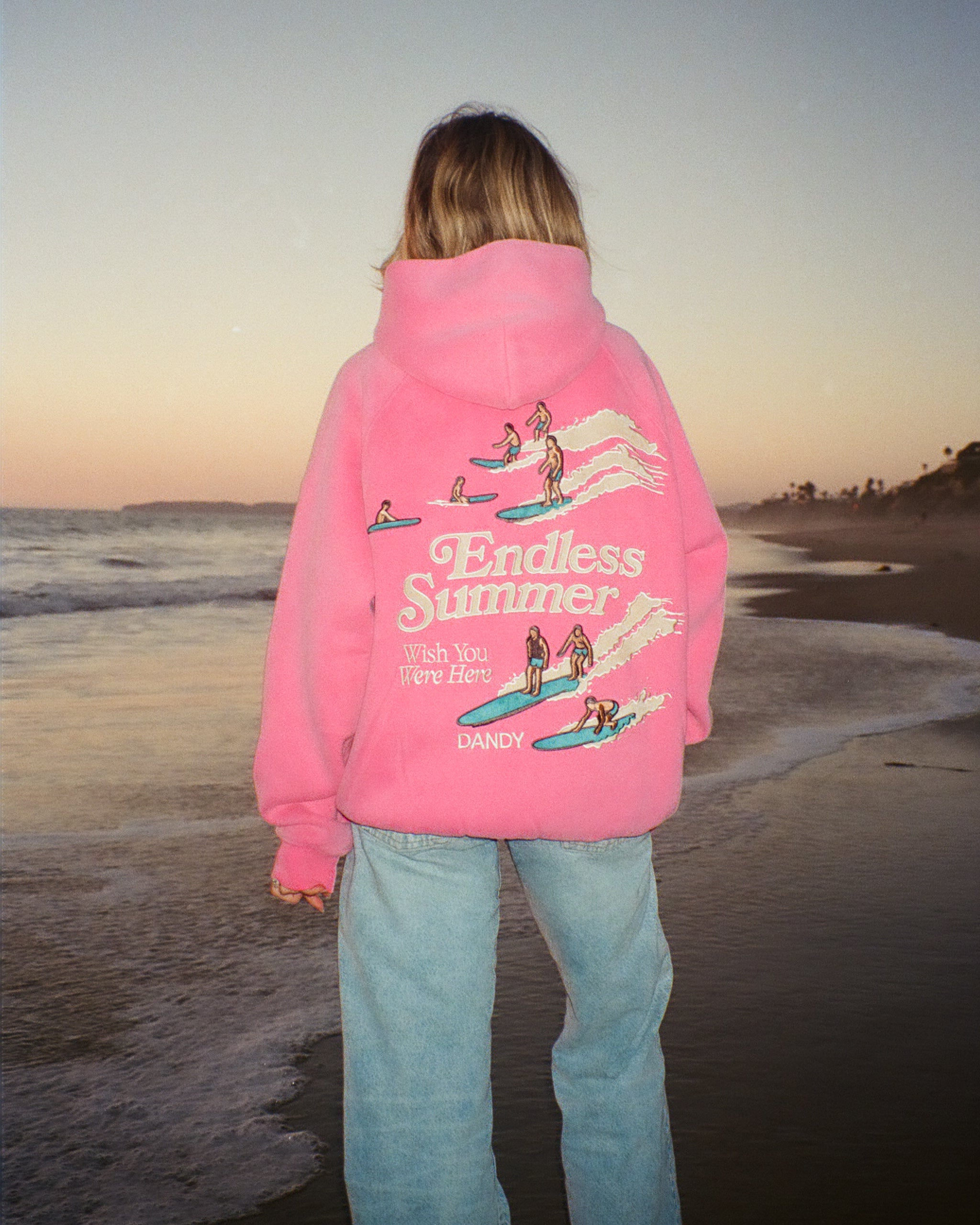 "Endless Summer" Oversized Lux Hoodie in Vintage Washed Pink standing on a sandy beach at sunset