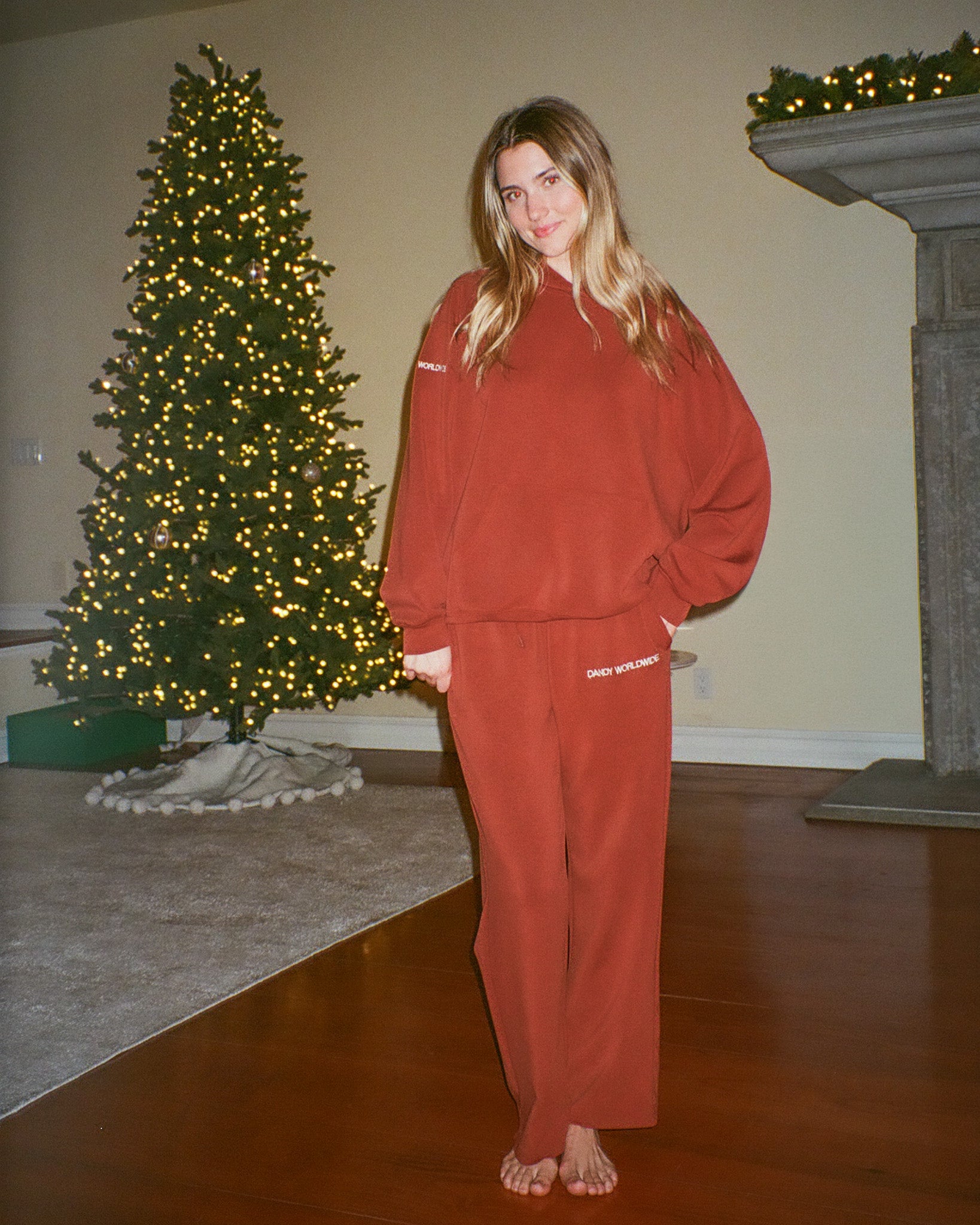 Person wearing a red outfit standing in a room with a decorated Christmas tree and fireplace.