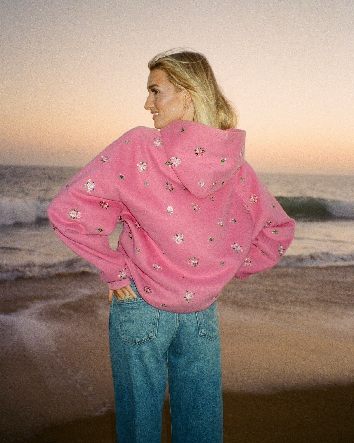 A woman stands on a beach at sunset wearing a pink oversized hoodie with small white floral prints and blue jeans, photographed from the back.