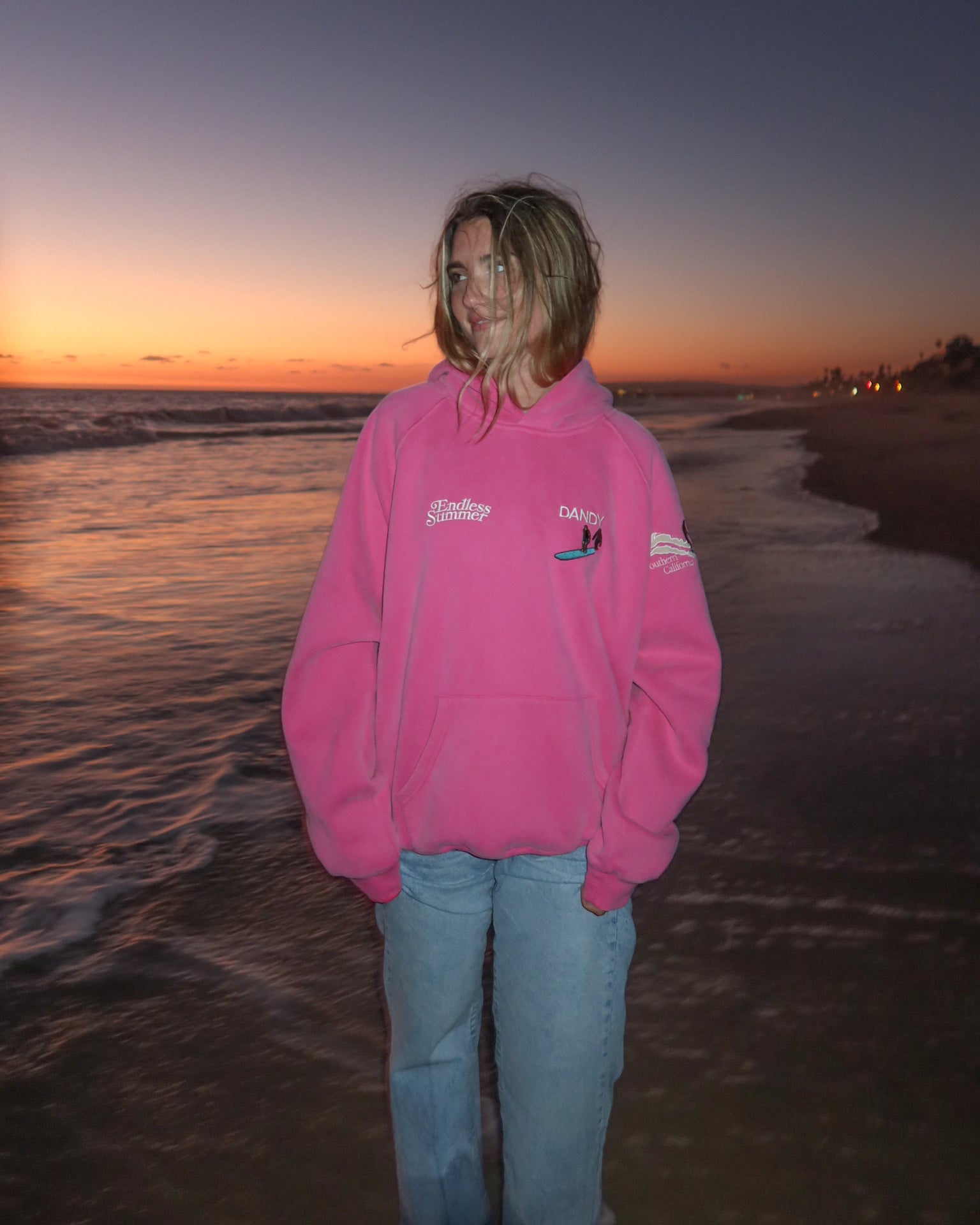 "Endless Summer" Oversized Lux Hoodie in Vintage Washed Pink standing on a sandy beach at sunset