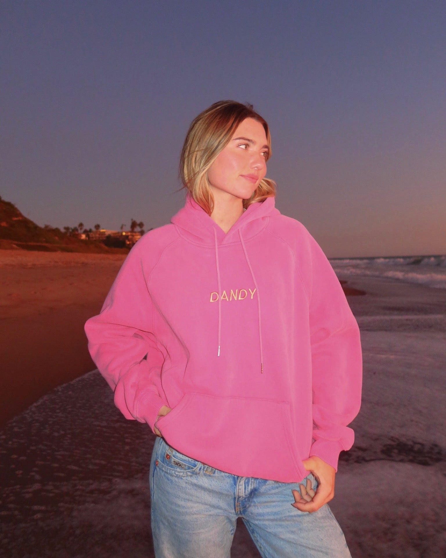 Person wearing a pink hoodie with 'DANDY' text on a beach at sunset.