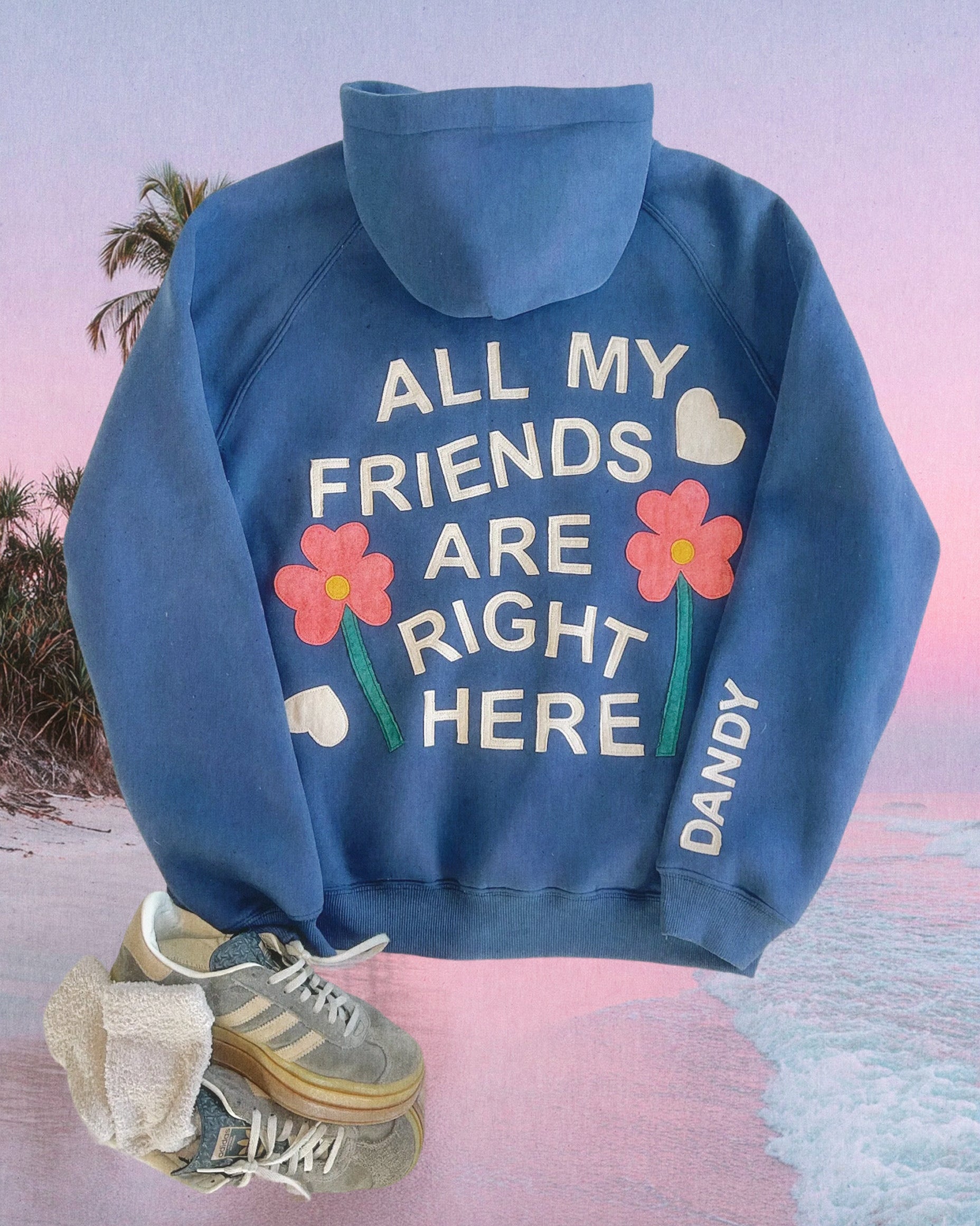 Photo shows a flat lay of a blue flower hoodie and gray Adidas against a graphic background of the beach.