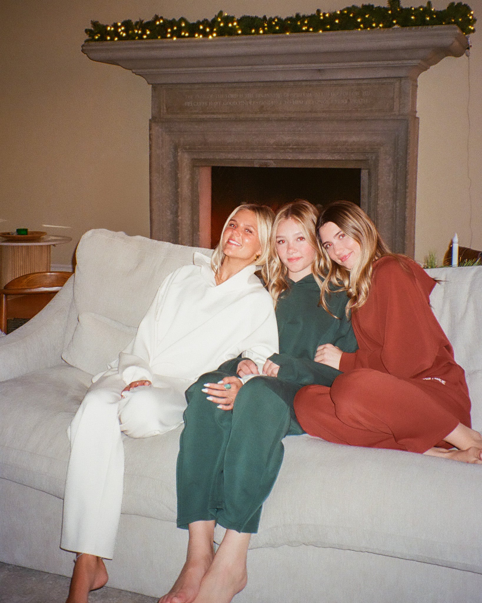 Three women sitting on a couch in front of a fireplace with Christmas decorations.