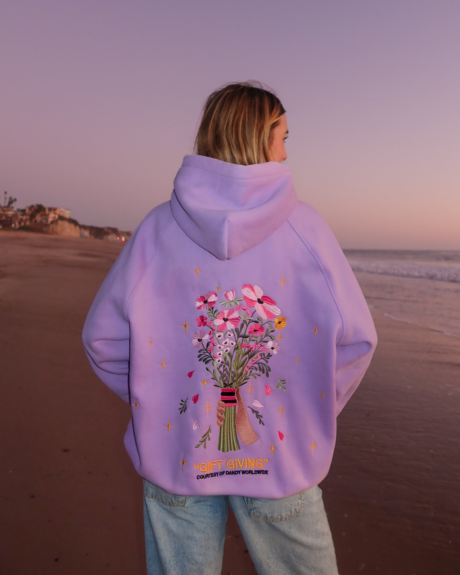 Person wearing a purple hoodie with floral design on a beach at sunset