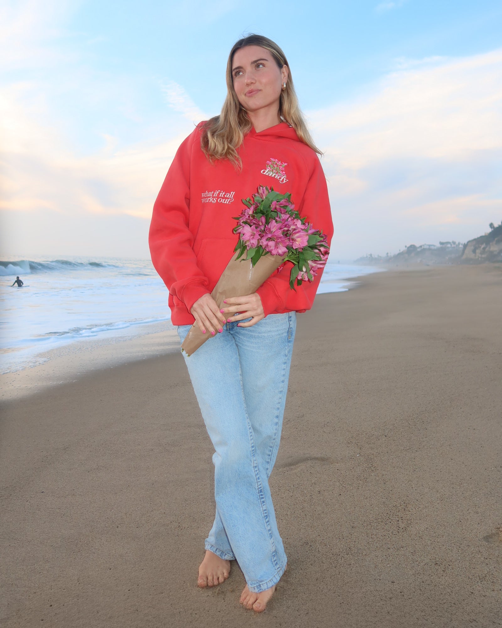 Woman in red sweatshirt and light blue jeans holding flowers on a beach