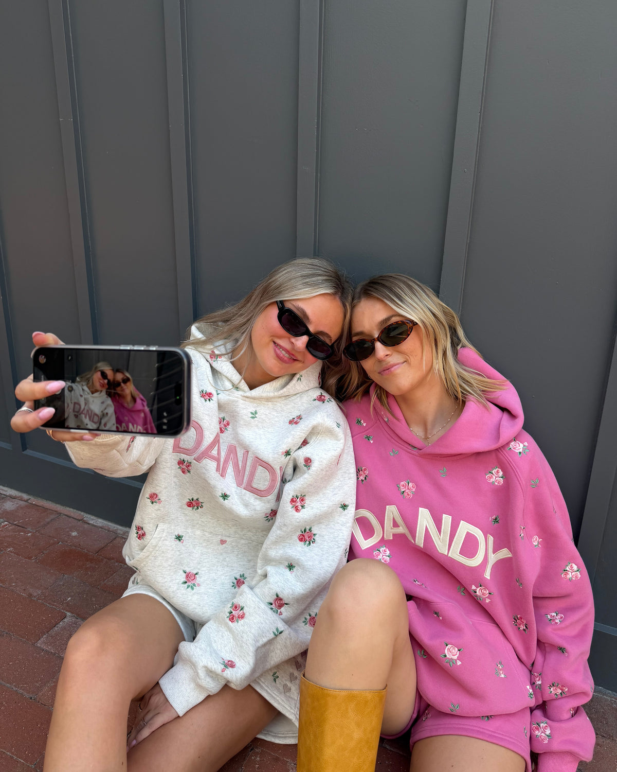 Two women taking a selfie outdoors against a dark gray paneled wall. One wears a cream hoodie with pink floral print and the word 'DANDY' in pink, while the other wears a bright pink hoodie with white floral print and the word 'DANDY' in white.