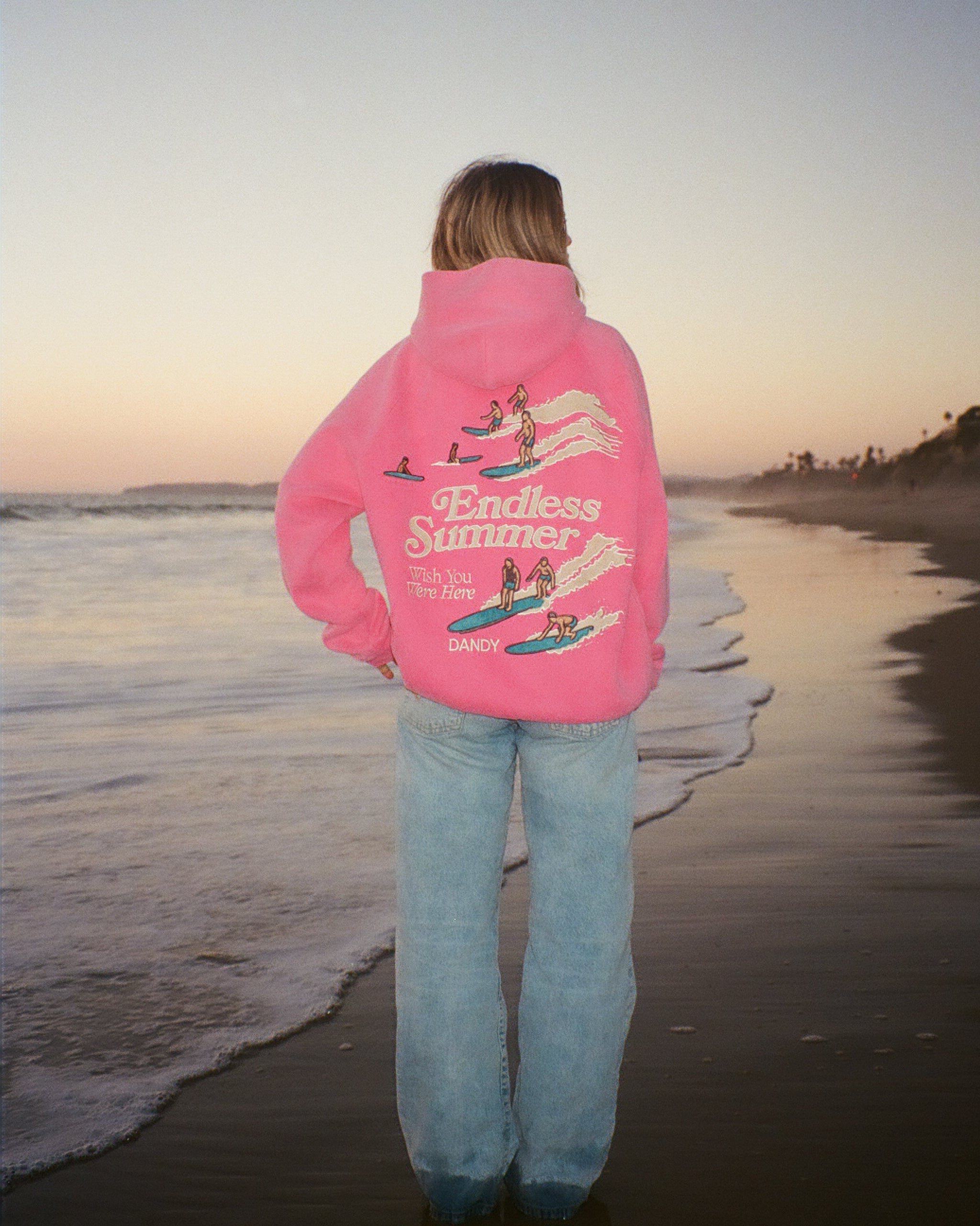 "Endless Summer" Oversized Lux Hoodie in Vintage Washed Pink standing on a sandy beach next to the water at sunset