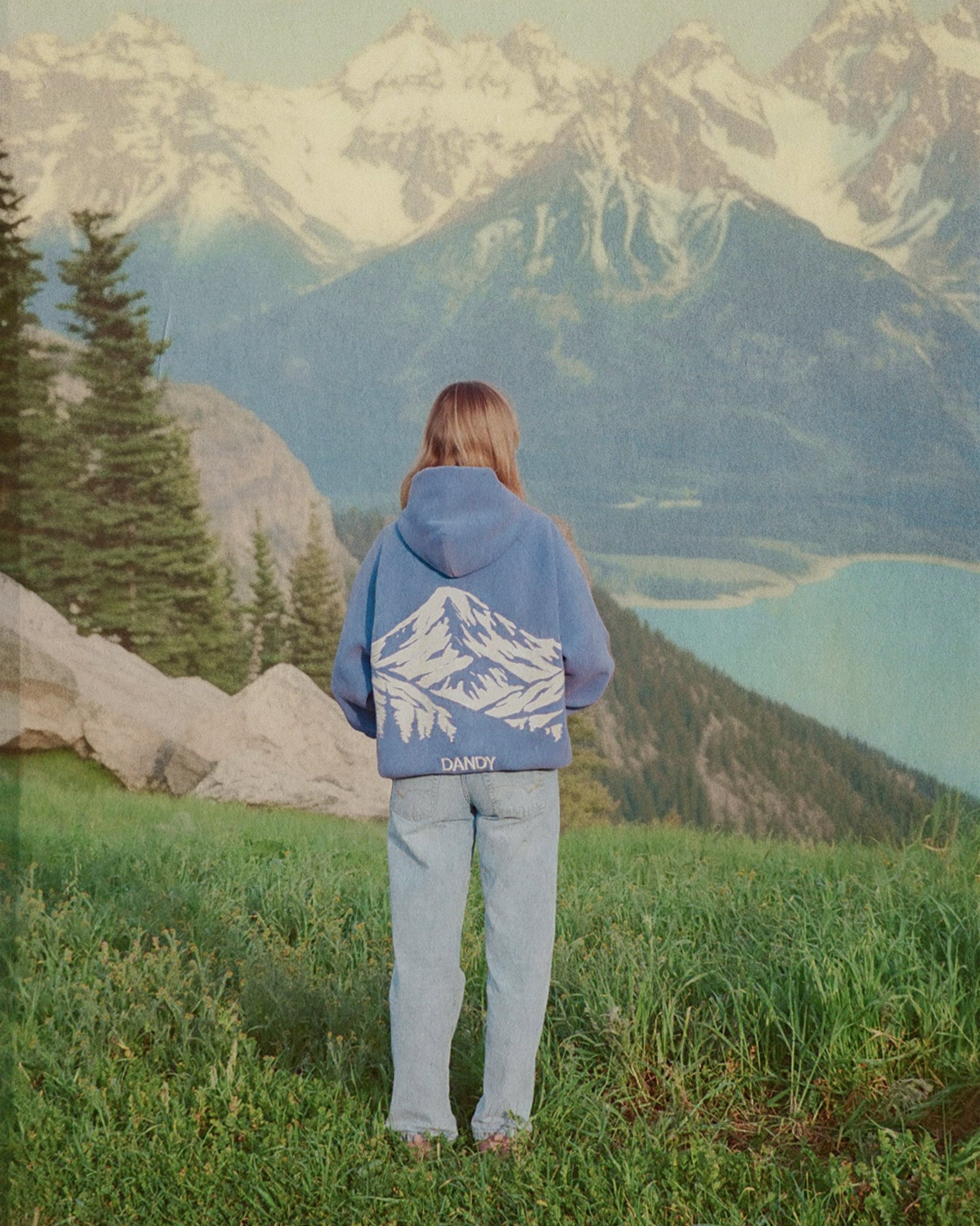 Person wearing a blue jacket with a mountain design, standing in front of a scenic view with mountains and a lake.