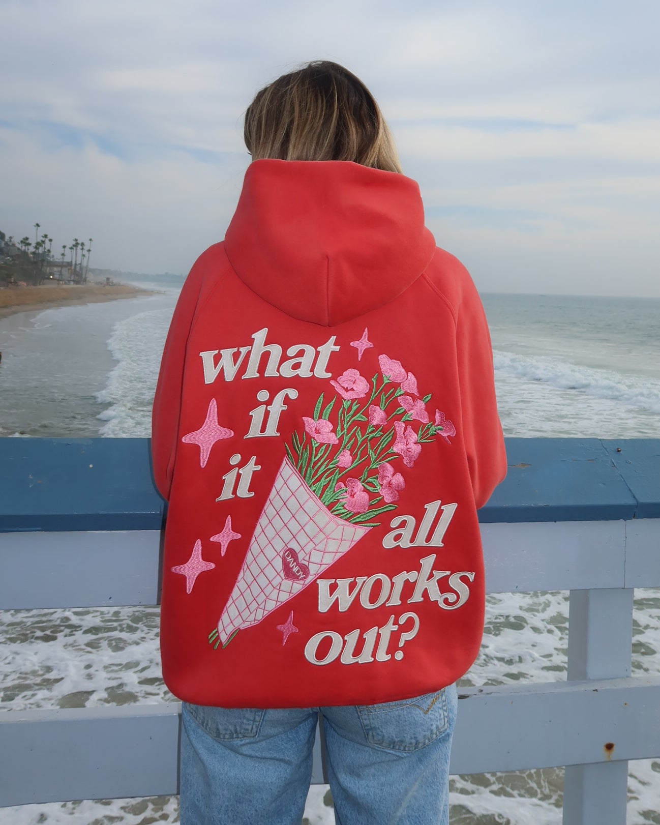 Person wearing a red hoodie with a floral design and text by the ocean.