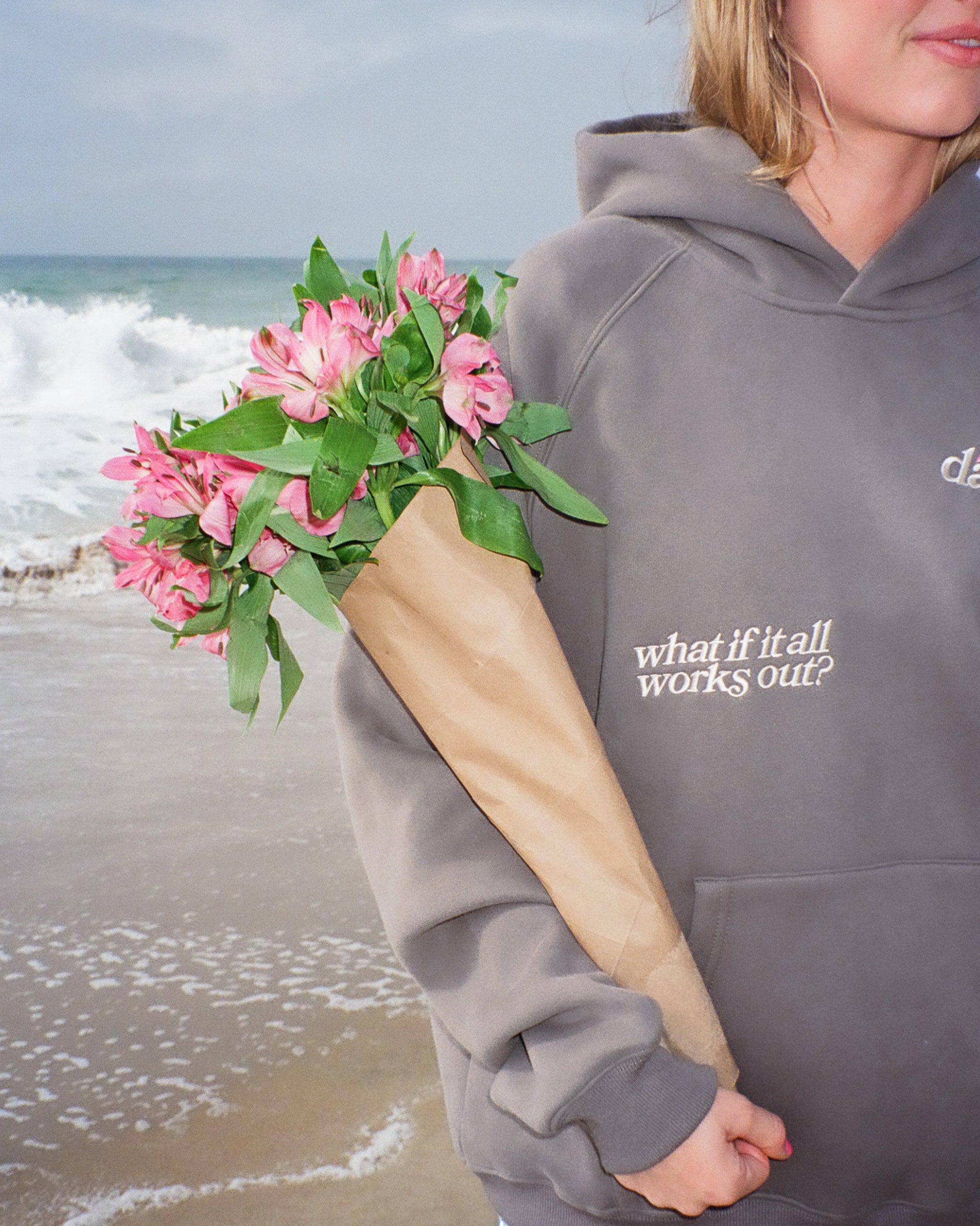 Person holding pink flowers with a hoodie that says 'what if it all works out?' on a beach.