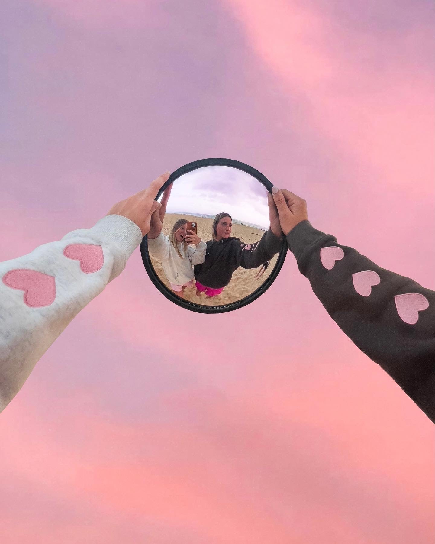 Two women hold circular mirror reflecting faces, in hoodies with pink heart sleeves, set against pastel pink sky