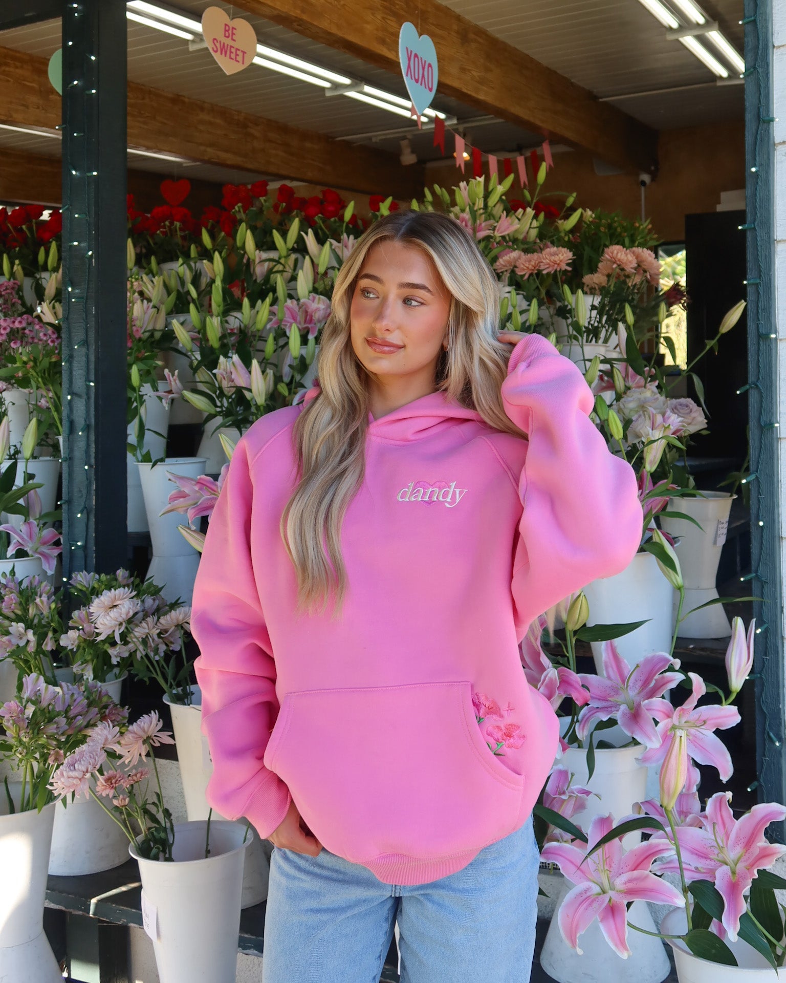 Woman wearing a pink hoodie with 'dandy' text in a flower shop.