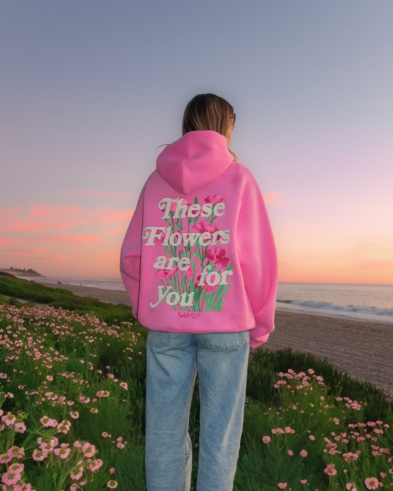Person wearing a pink hoodie with text standing in a field of flowers at sunset.