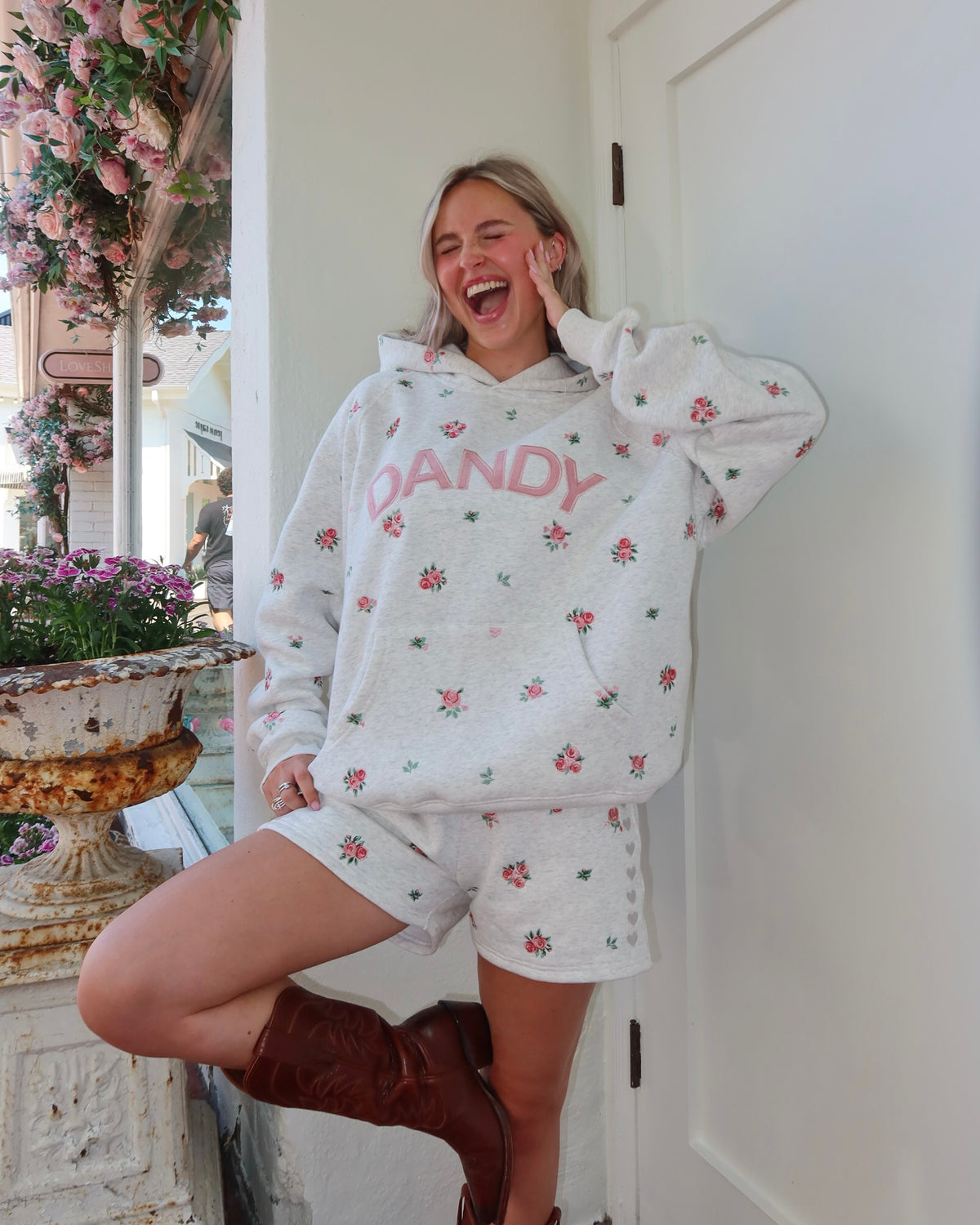 A woman wearing a white heathered hoodie and matching shorts set with pink floral prints and the word 'DANDY' in pink on the chest, posing on a porch next to a white door.
