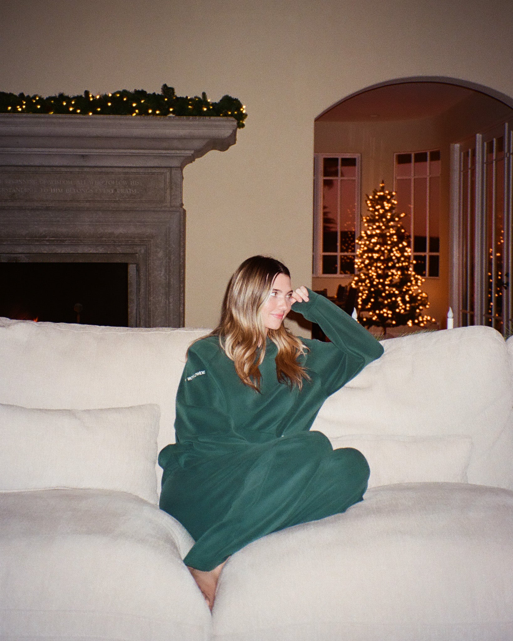 Woman in green matching set sitting on a couch with a Christmas tree and fireplace in the background