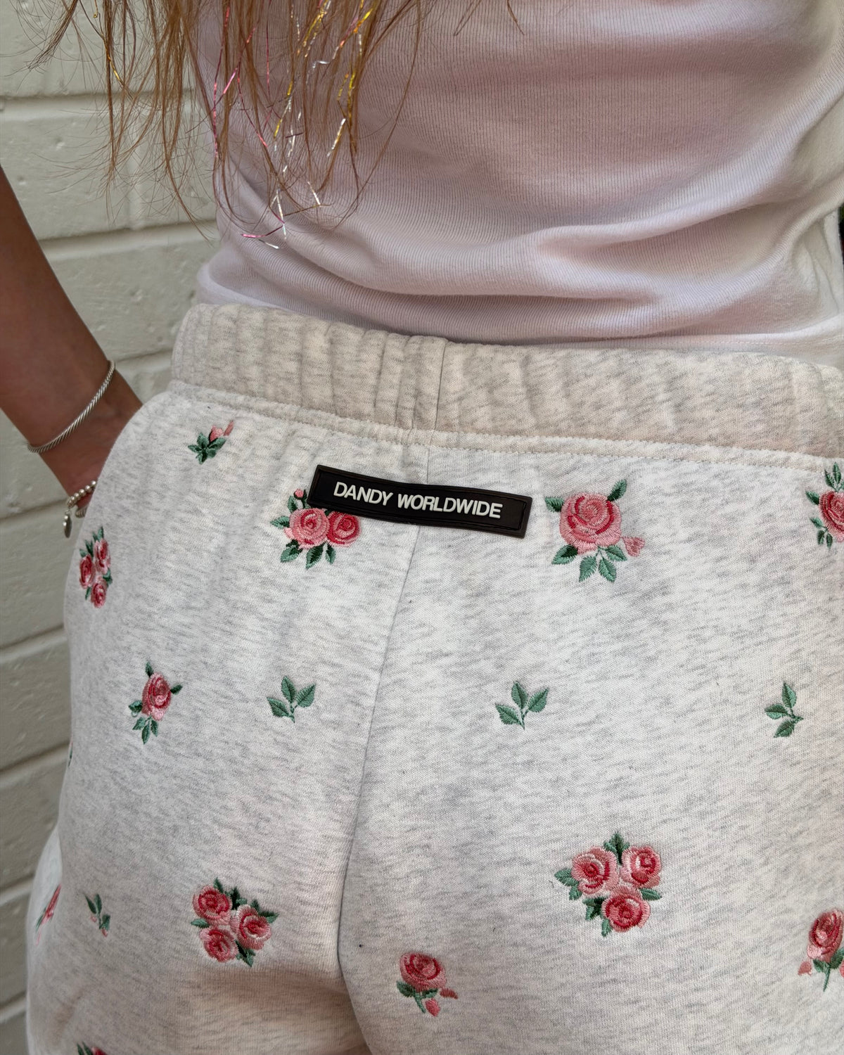 Close-up of a person wearing light heather gray sweatpants with a pink and green rose pattern and a black 'DANDY WORLDWIDE' label at the back waistband, paired with a pale pink top.