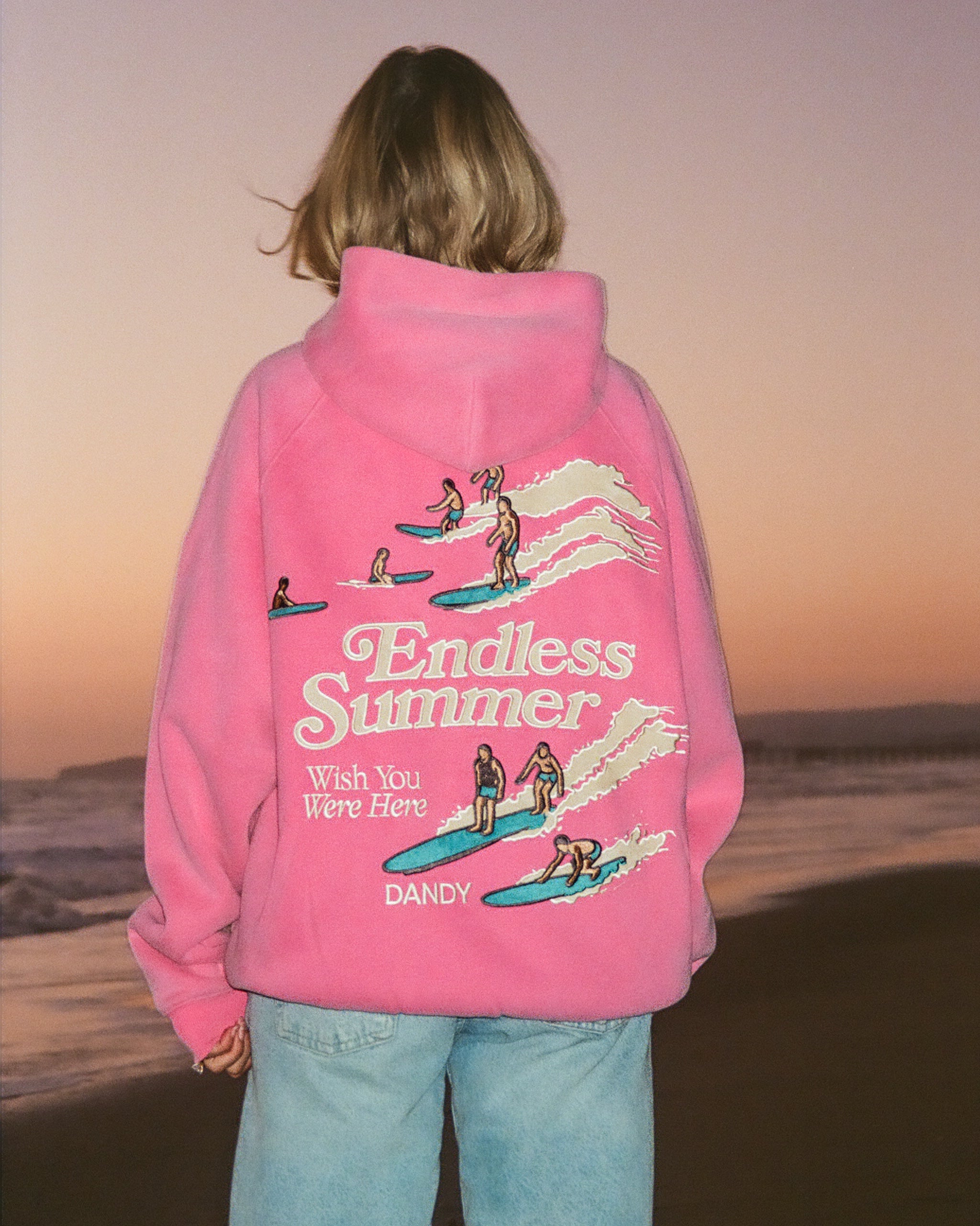 "Endless Summer" Oversized Lux Hoodie in Vintage Washed Pink standing on a sandy beach at sunset