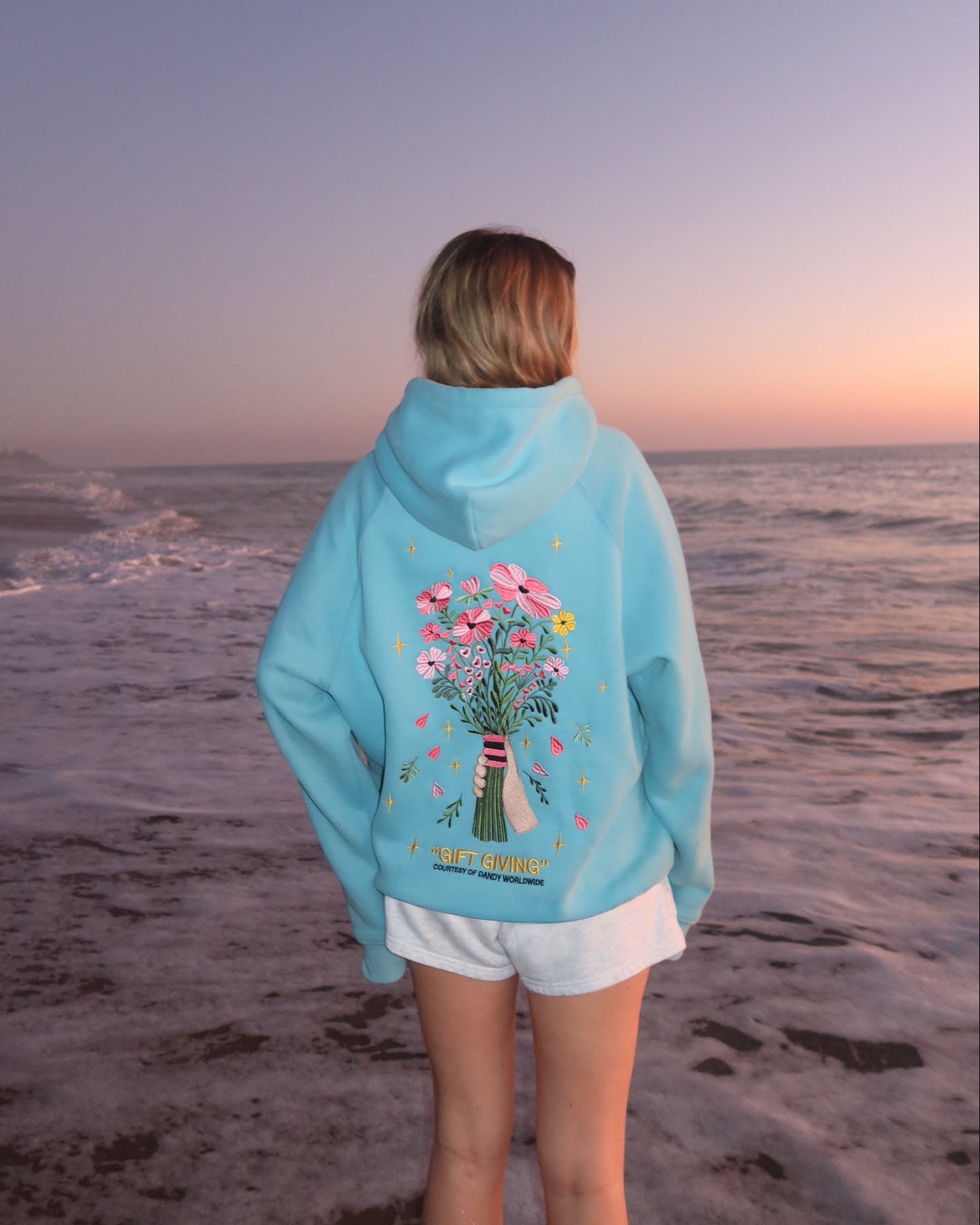 Person wearing a blue hoodie with a floral design on the beach at sunset