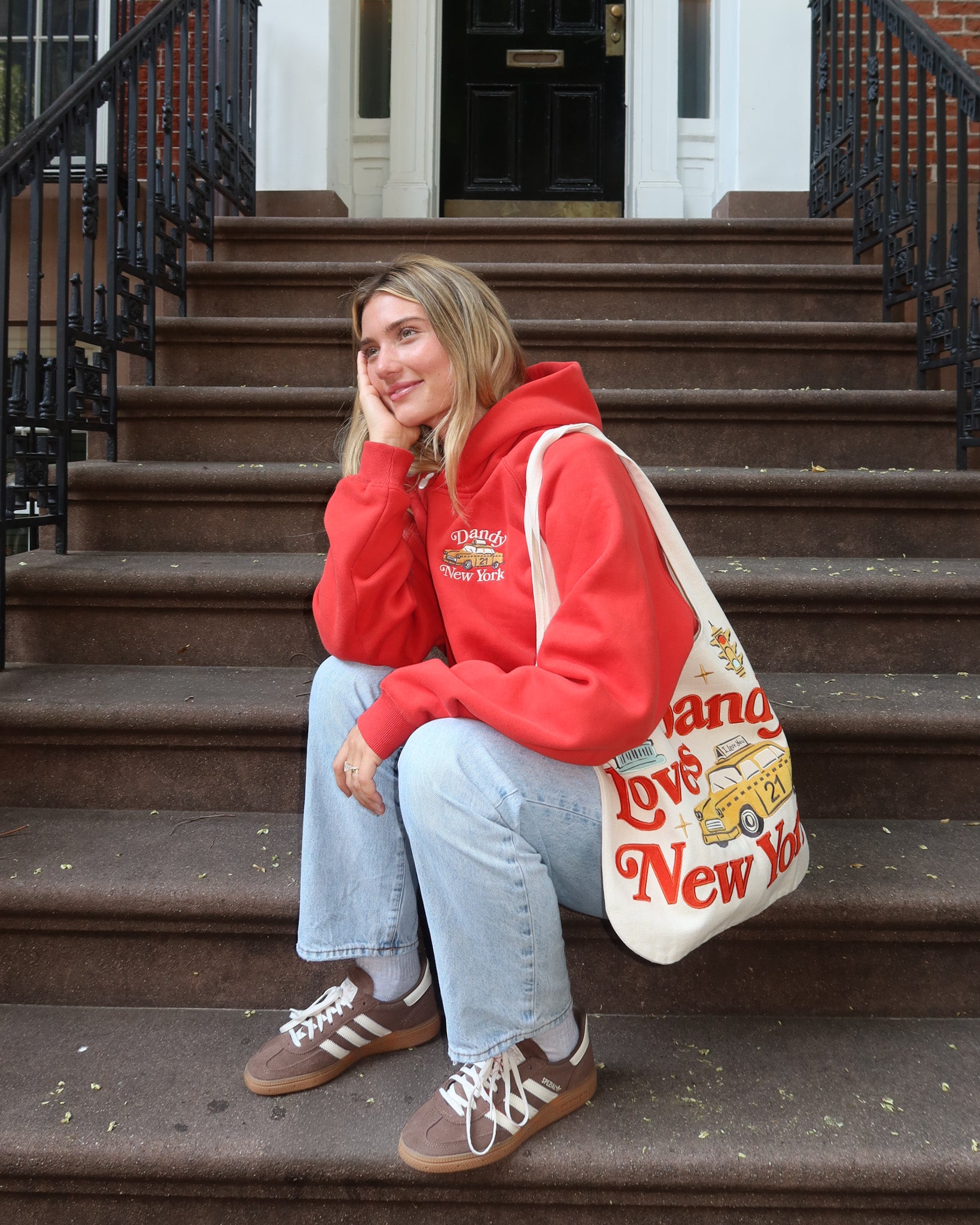 Person sitting on steps wearing a red hoodie and blue jeans, holding a 'Dandy Loves New York' bag.