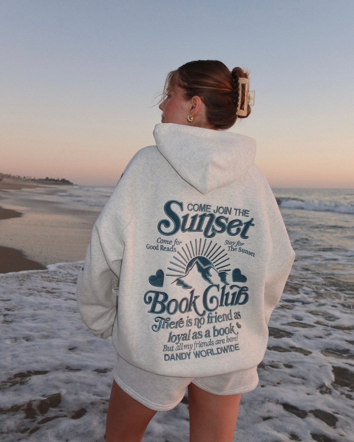 Person wearing a hoodie with a sunset book club design on a beach at sunset.