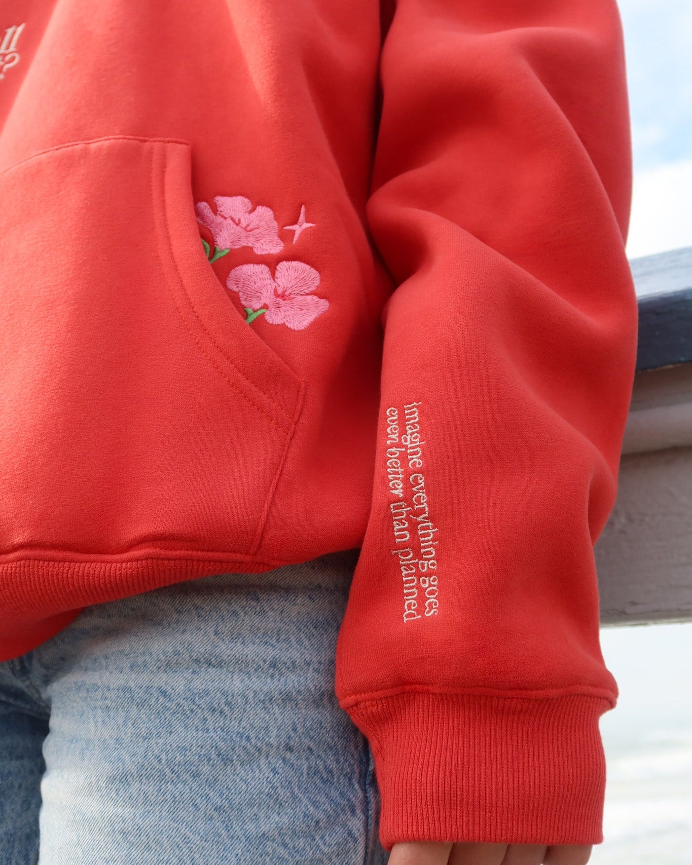 Red hoodie with floral design and text on sleeve, worn by a person.