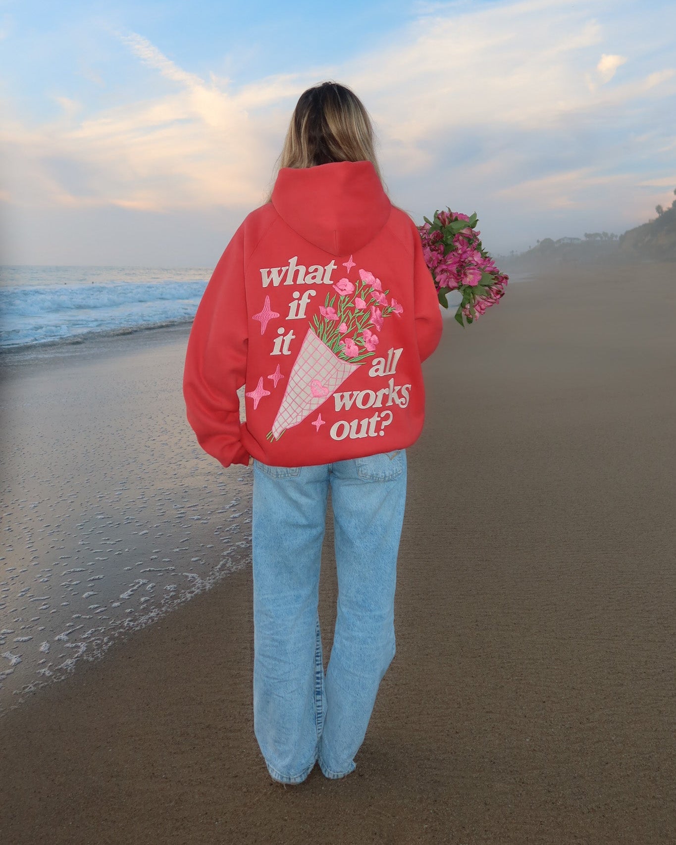 Person on a beach wearing a red jacket with text and graphics, holding flowers.