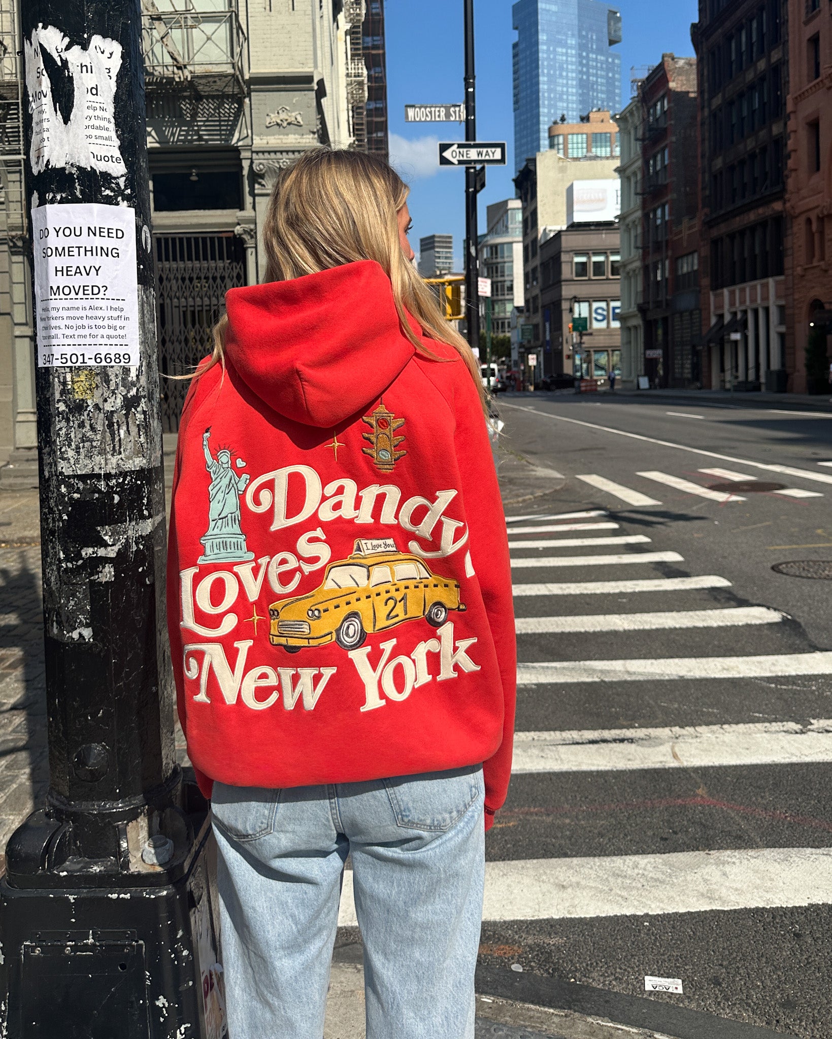 Person wearing a red hoodie with 'Dandy Loves New York' text and taxi graphic on a city street.