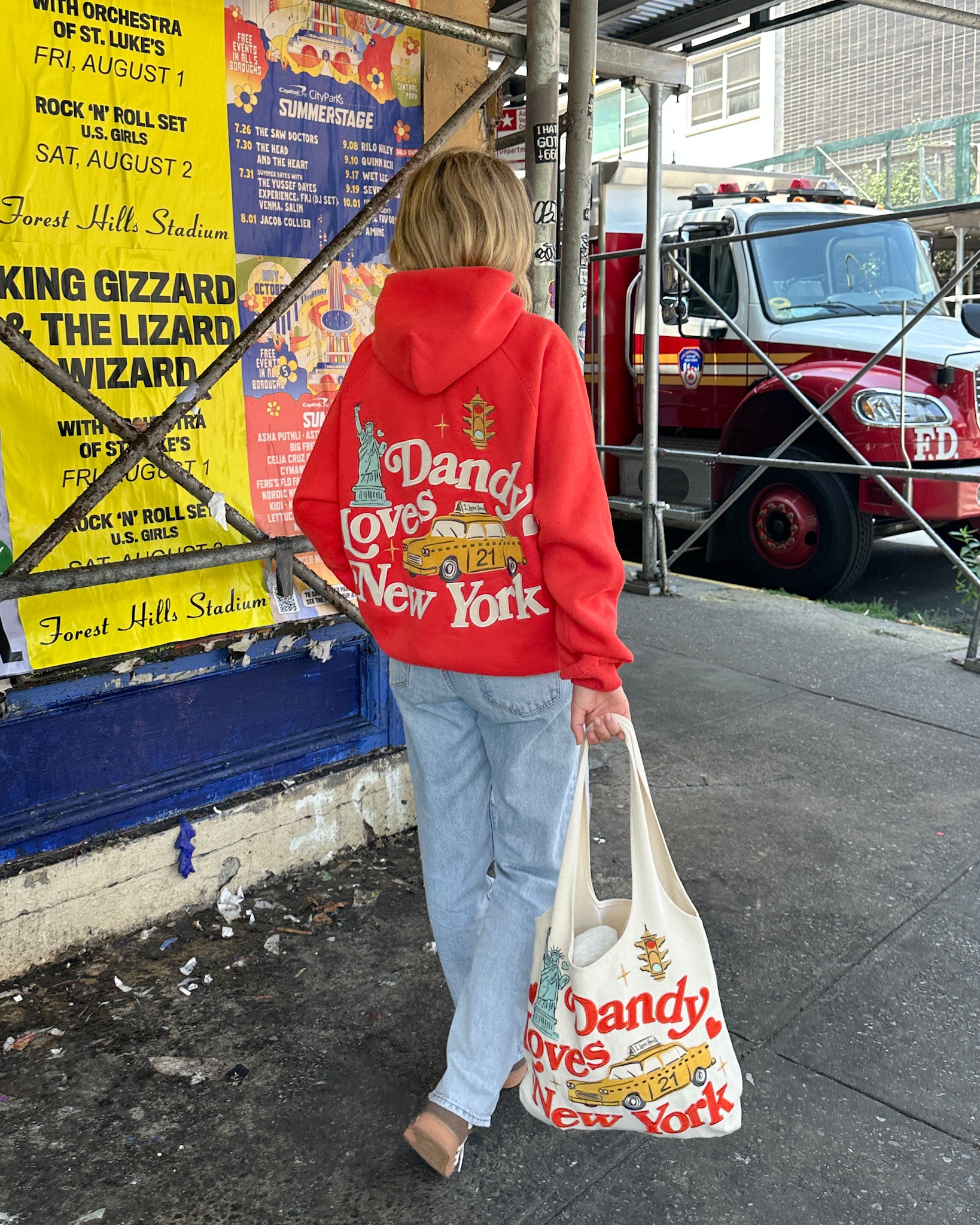 Person wearing a red hoodie with 'Dandy Loves New York' design, holding a matching tote bag, walking past a fire truck and concert posters.