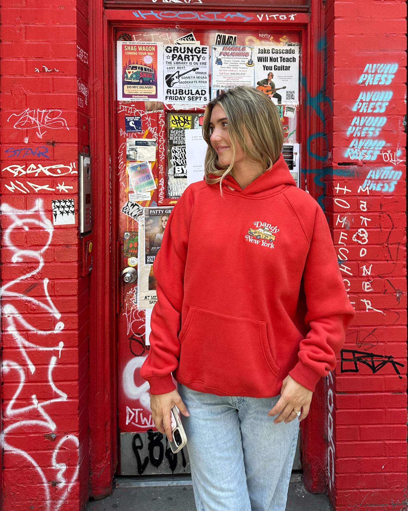 Person wearing a red hoodie in front of a red door with graffiti and posters.