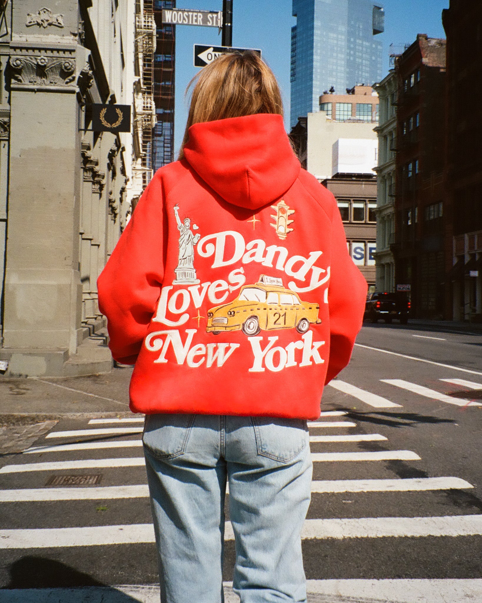 Person wearing a red hoodie with 'Dandy Loves New York' text and graphics, standing on a city street.