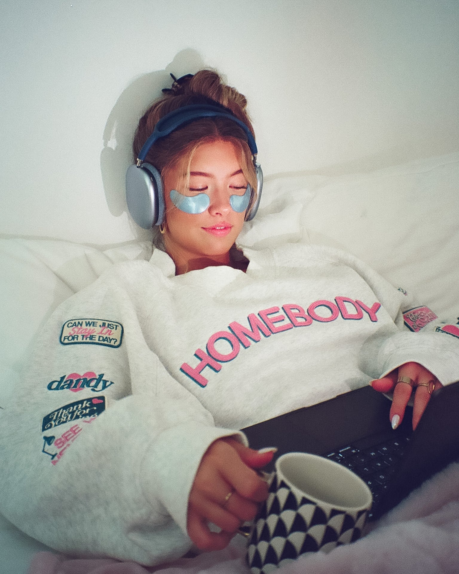 Person wearing a 'Homebody' sweatshirt with patches, lying on a bed with a mug and headphones.