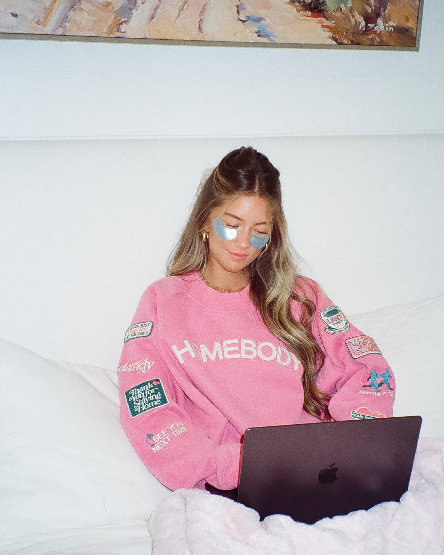 Person wearing a pink 'Homebody' sweatshirt with patches, using a laptop on a bed.