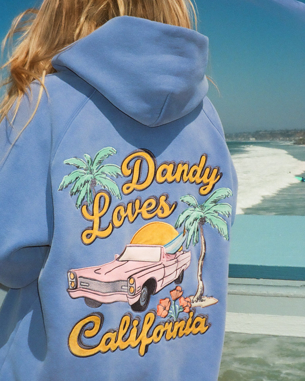 Person wearing a blue hoodie with 'Dandy Loves California' design by the beach.