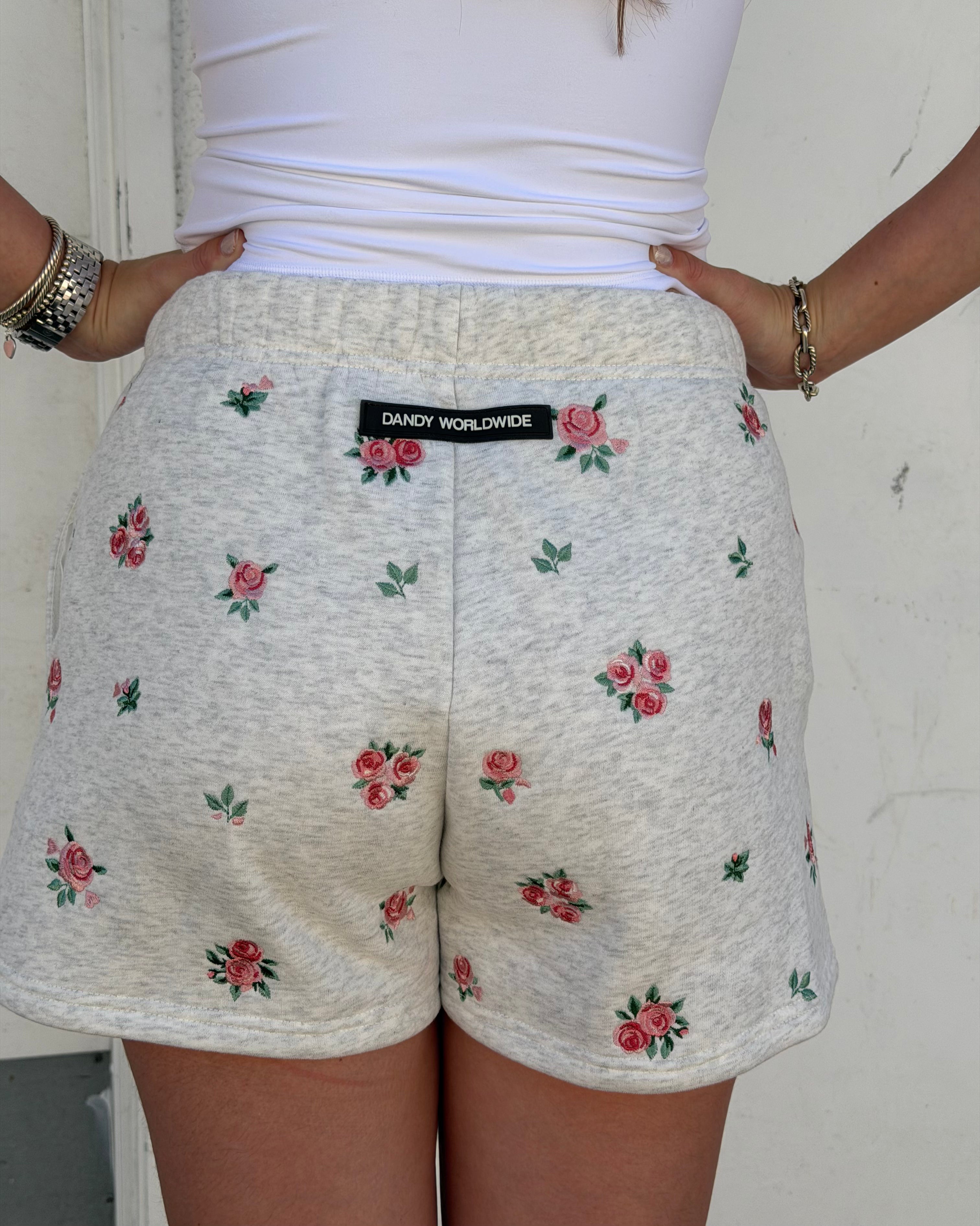 A person wearing light heather gray shorts with a pink and green rose pattern, paired with a white top. The shorts feature a black waistband label that reads 'DANDY WORLDWIDE'.