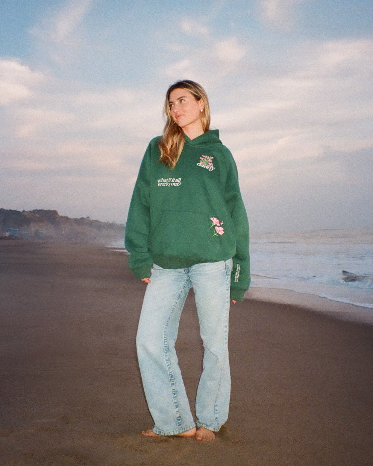 Person wearing a green hoodie and light blue jeans standing on a beach with ocean and sky in the background.