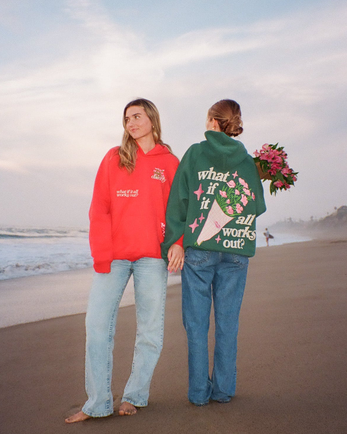 Two women standing on a beach, one in a red sweatshirt and light blue jeans, the other in a green sweatshirt with floral design and blue jeans, holding flowers.
