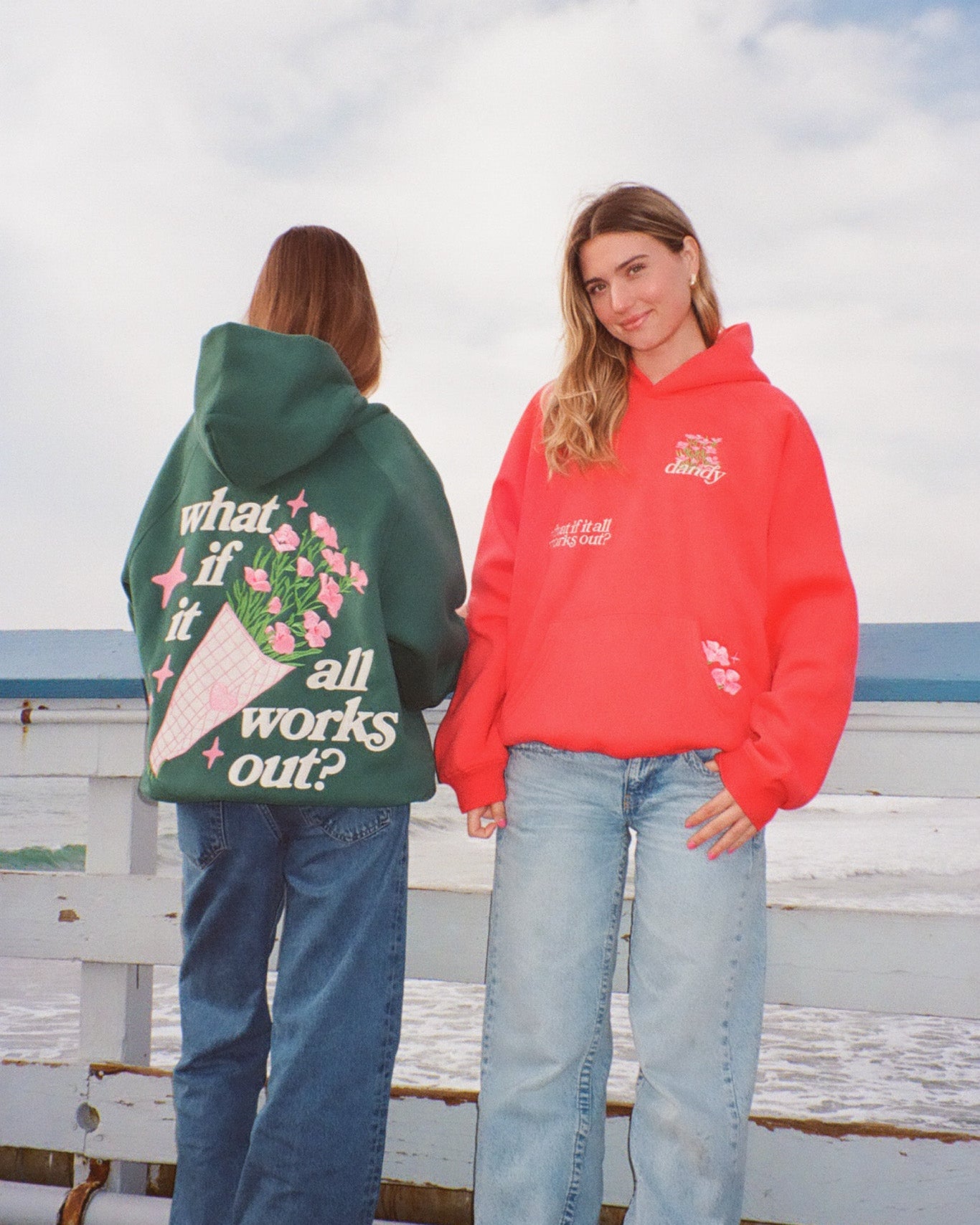 Two people wearing hoodies with printed designs by a beach.