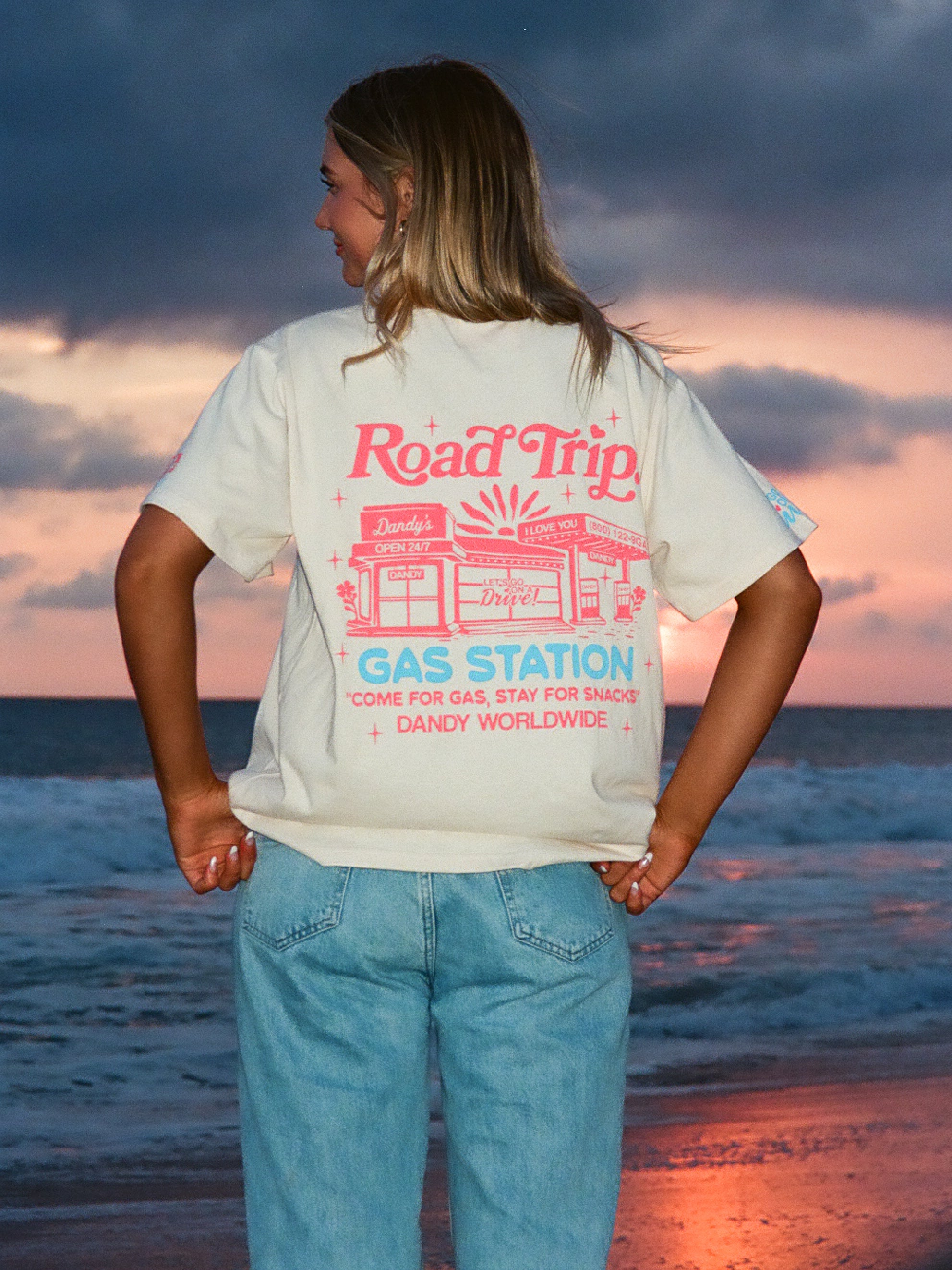 Person wearing a t-shirt with 'Road Trip Gas Station' design on a beach at sunset.