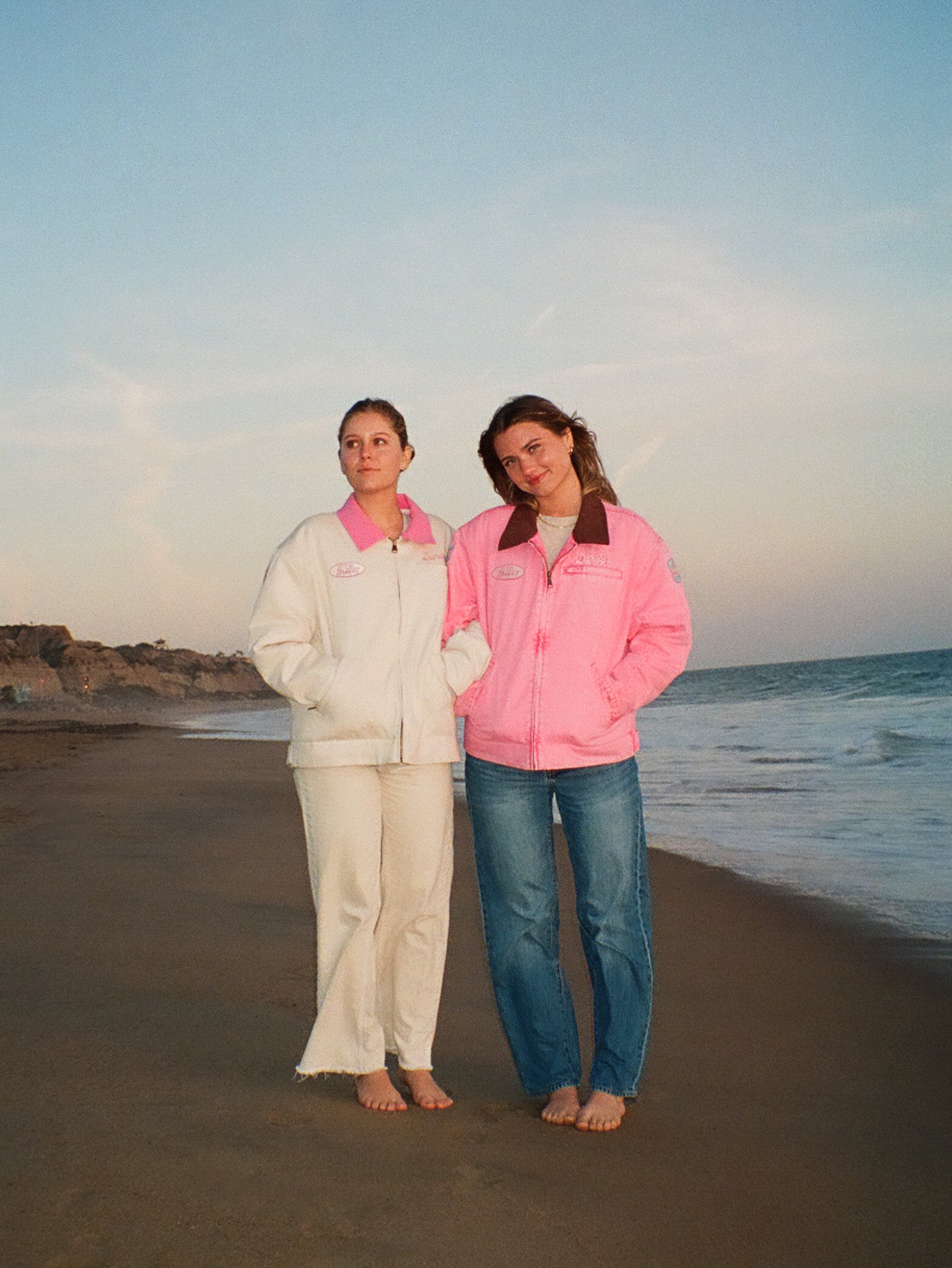 Two women standing on a beach wearing matching pink and white outfits.